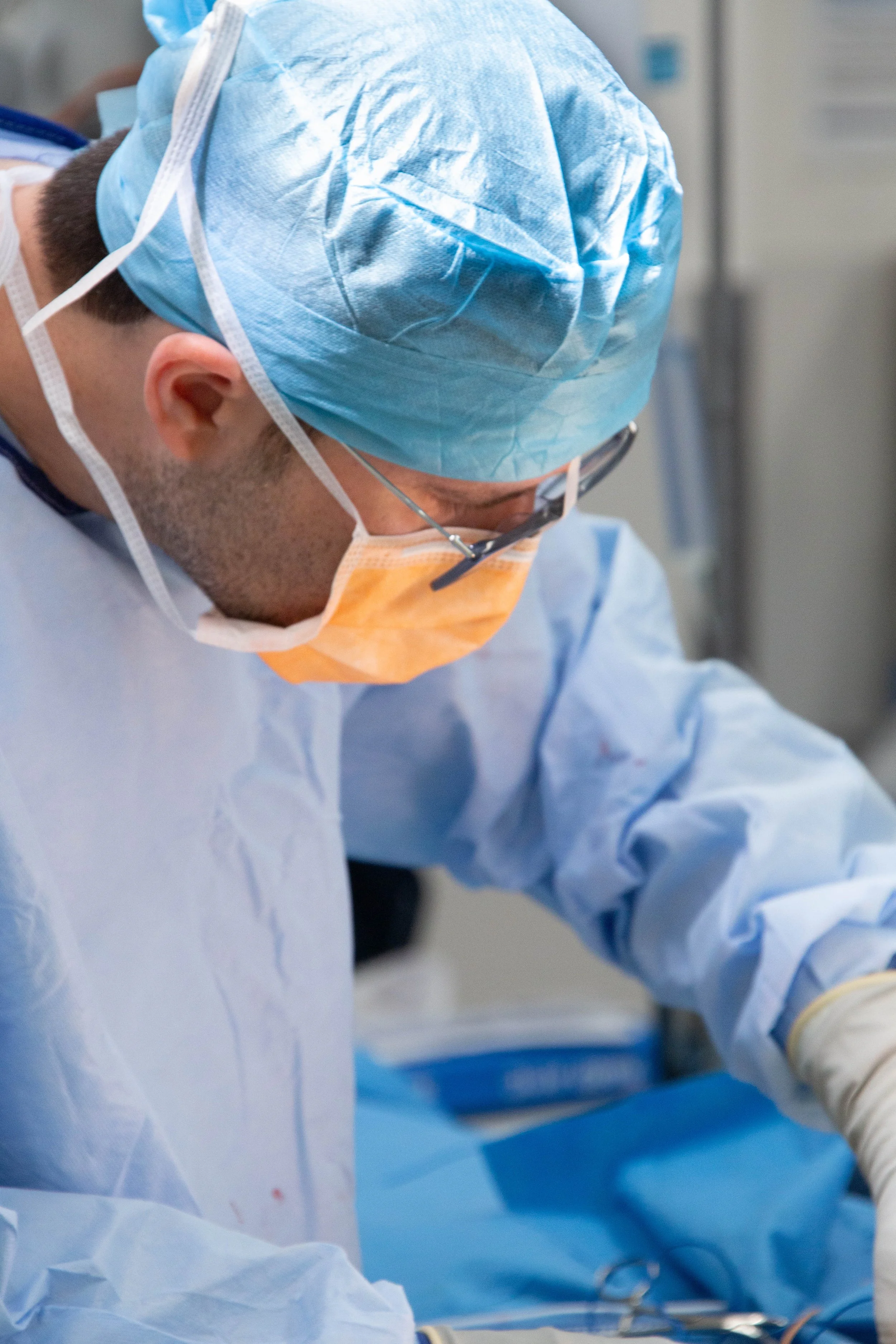 A surgeon dressed in blue surgical scrubs, a blue surgical cap, glasses, and a face mask, likely performing or assisting in a surgical procedure