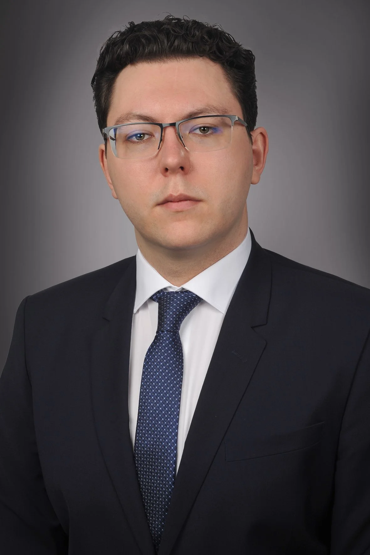 Portrait of a man with short dark hair, glasses, wearing a black suit, white dress shirt, and blue patterned tie, against a gray background.
