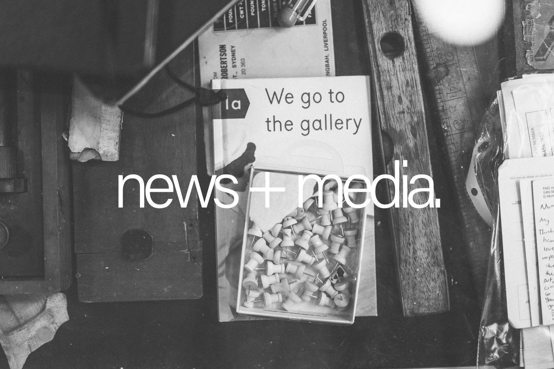 A close-up view of a cluttered wooden table with various objects on it. There is a box of numerous small white objects, possibly thumbtacks or push pins. A sign that says "We go to the gallery" is partially visible, along with papers, a container, and some other miscellaneous items. The photo appears to be in black and white.