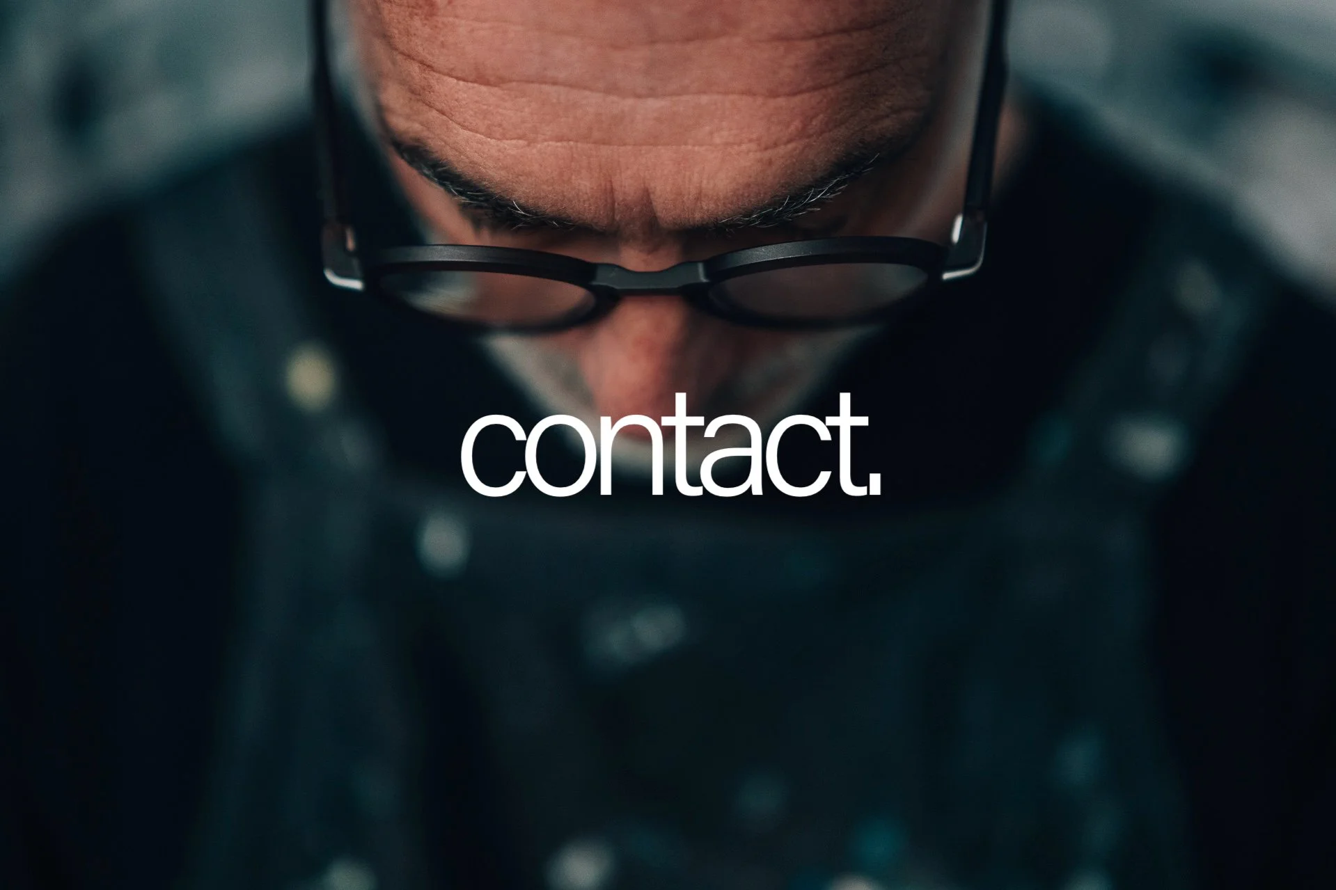 Close-up of a person's face looking down, wearing black glasses, with a focus on the forehead and eyebrows. The background is blurred, and the word 'contact.' is overlaid in white text at the bottom of the image.