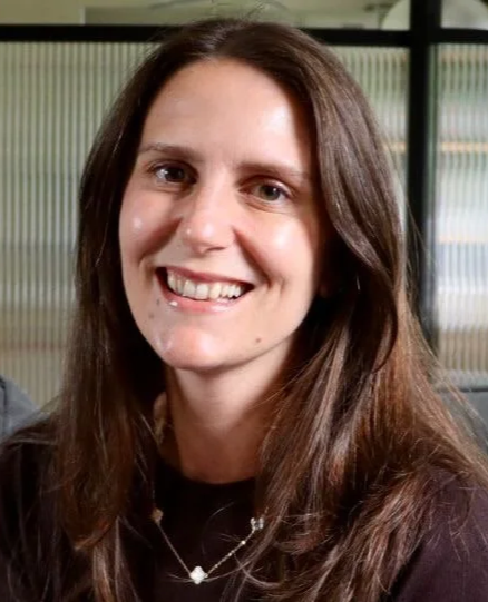 A woman with long brown hair smiling in an indoor setting with glass windows in the background.