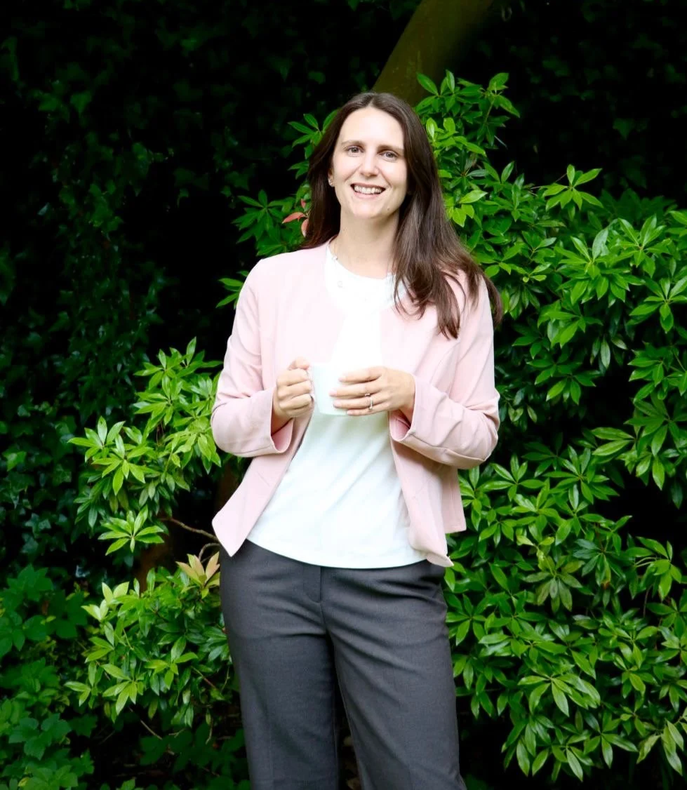A woman with long brown hair, smiling, holding a white mug, standing outdoors in front of green leafy bushes, wearing a pink blazer over a white top and dark gray pants.