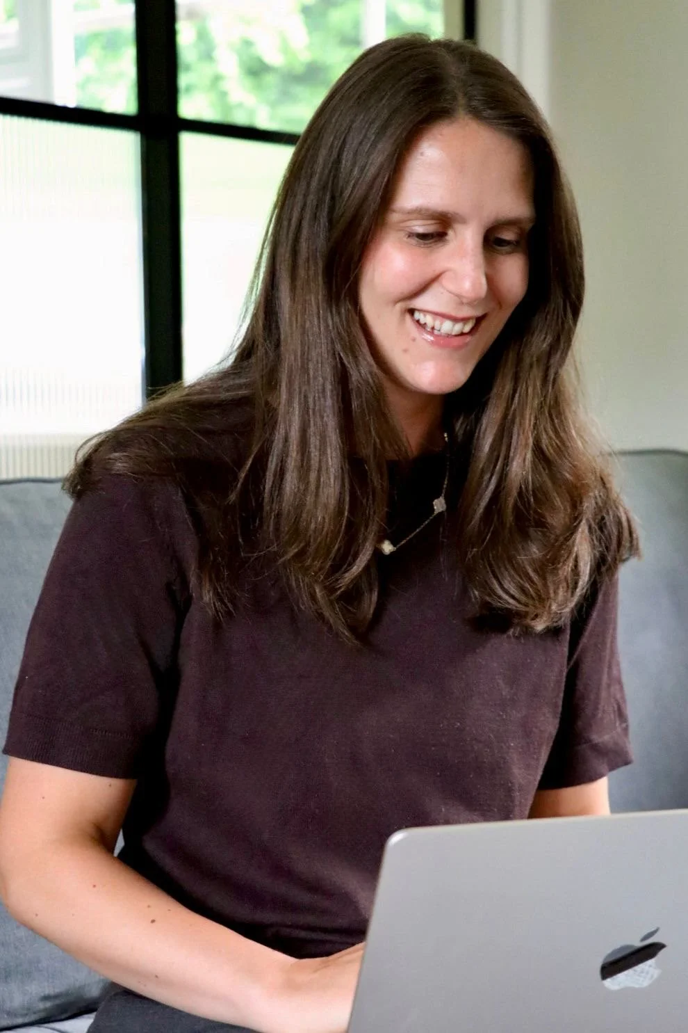 A woman with long brown hair wearing a dark T-shirt, smiling while looking at a laptop.