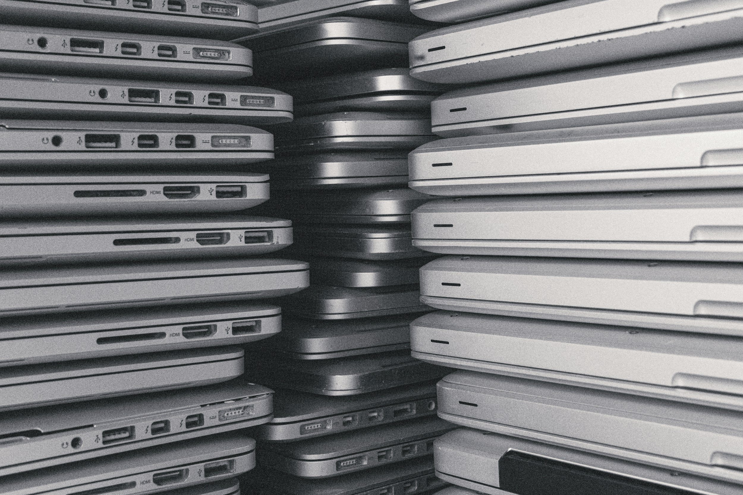 Stacked laptops in various shades of gray and white, viewed from the side.