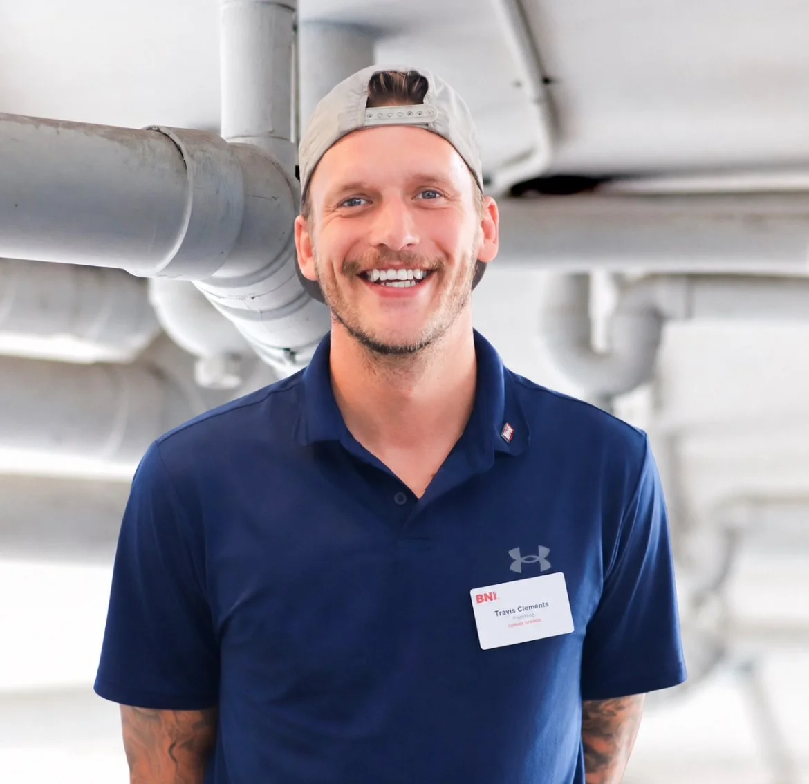 Smiling man wearing a navy polo shirt, gray baseball cap backward, and a name tag standing under industrial pipes.