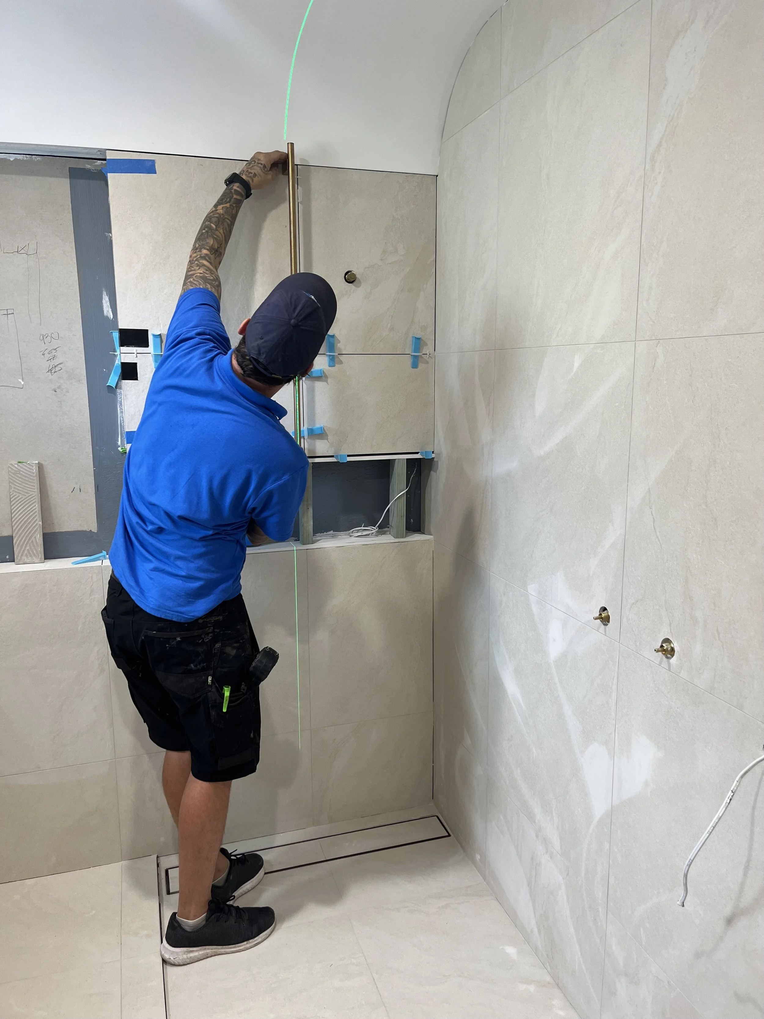 A man in a blue t-shirt, black shorts, and a black cap is using a laser level to align tiles on a wall in a bathroom. The bathroom has beige tiled walls and floors, with visible plumbing fixtures.