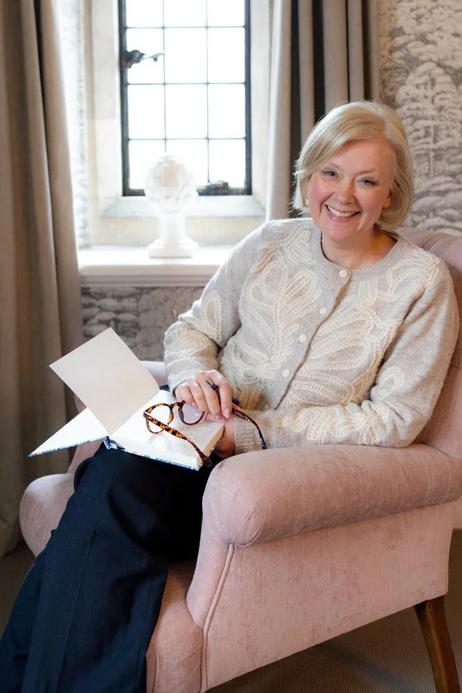 A woman with blonde hair smiling and sitting in a pink armchair in a room with a window, curtains, and a decorative vase on the windowsill, holding glasses and a notebook.