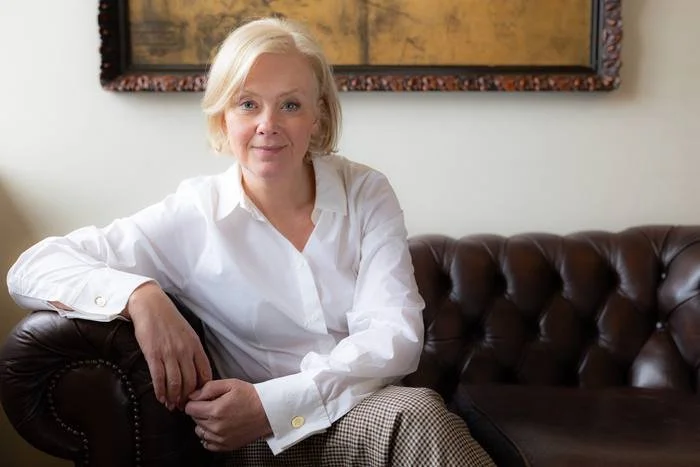 A woman with blonde hair sits on a brown leather couch in a living room, wearing a white shirt and beige checkered pants, with a neutral expression. Behind her, there is a painting on the wall.