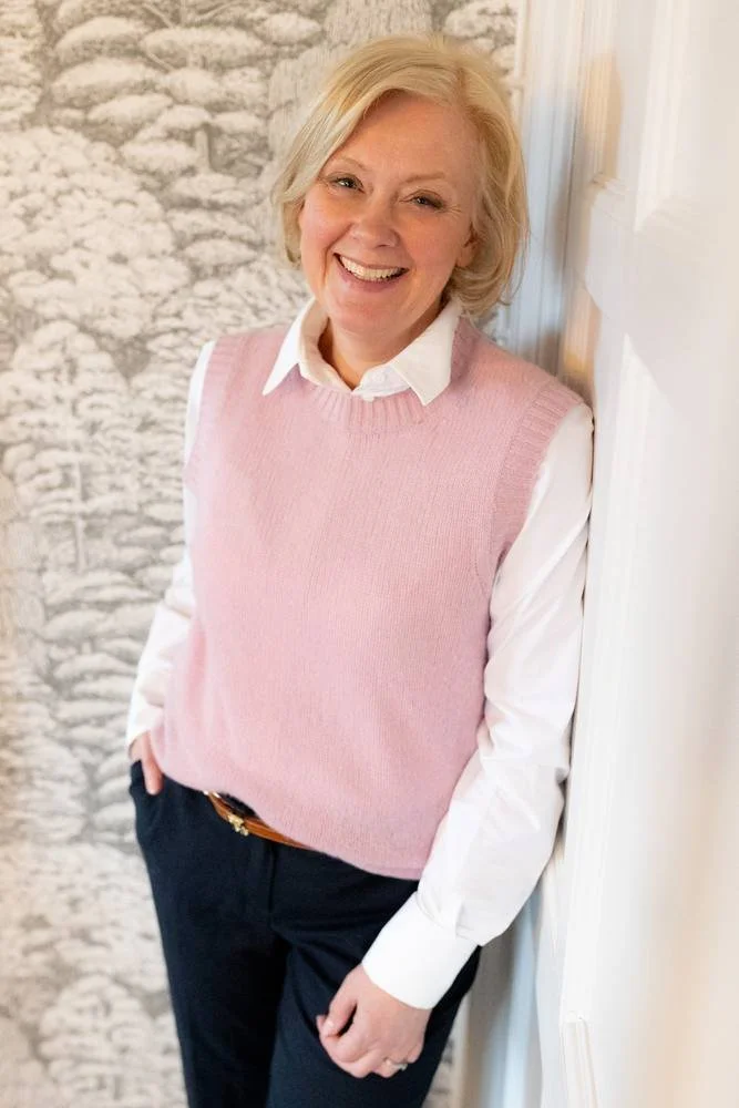 A smiling blonde woman dressed in a pink vest over a white shirt, standing indoors near a corner with a stone wall and a white door.