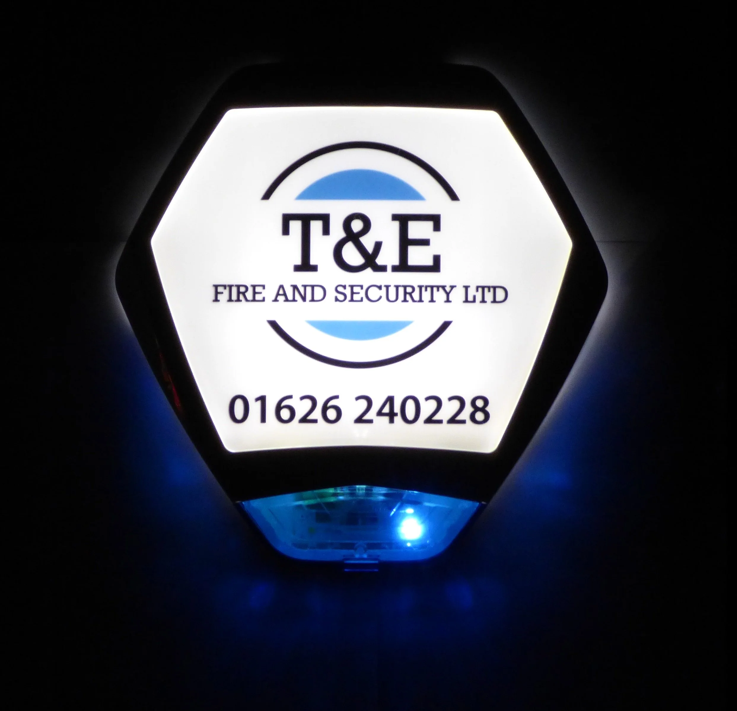 Close-up of a Fire & Security Ltd logo on a glowing white hexagon-shaped sign, with black text and blue design elements, emitting blue light in a dark environment.