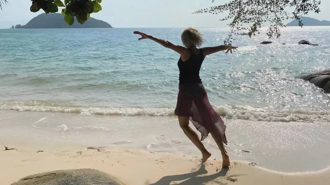 Person dancing on a sandy beach near the water, with mountains and trees in the background.