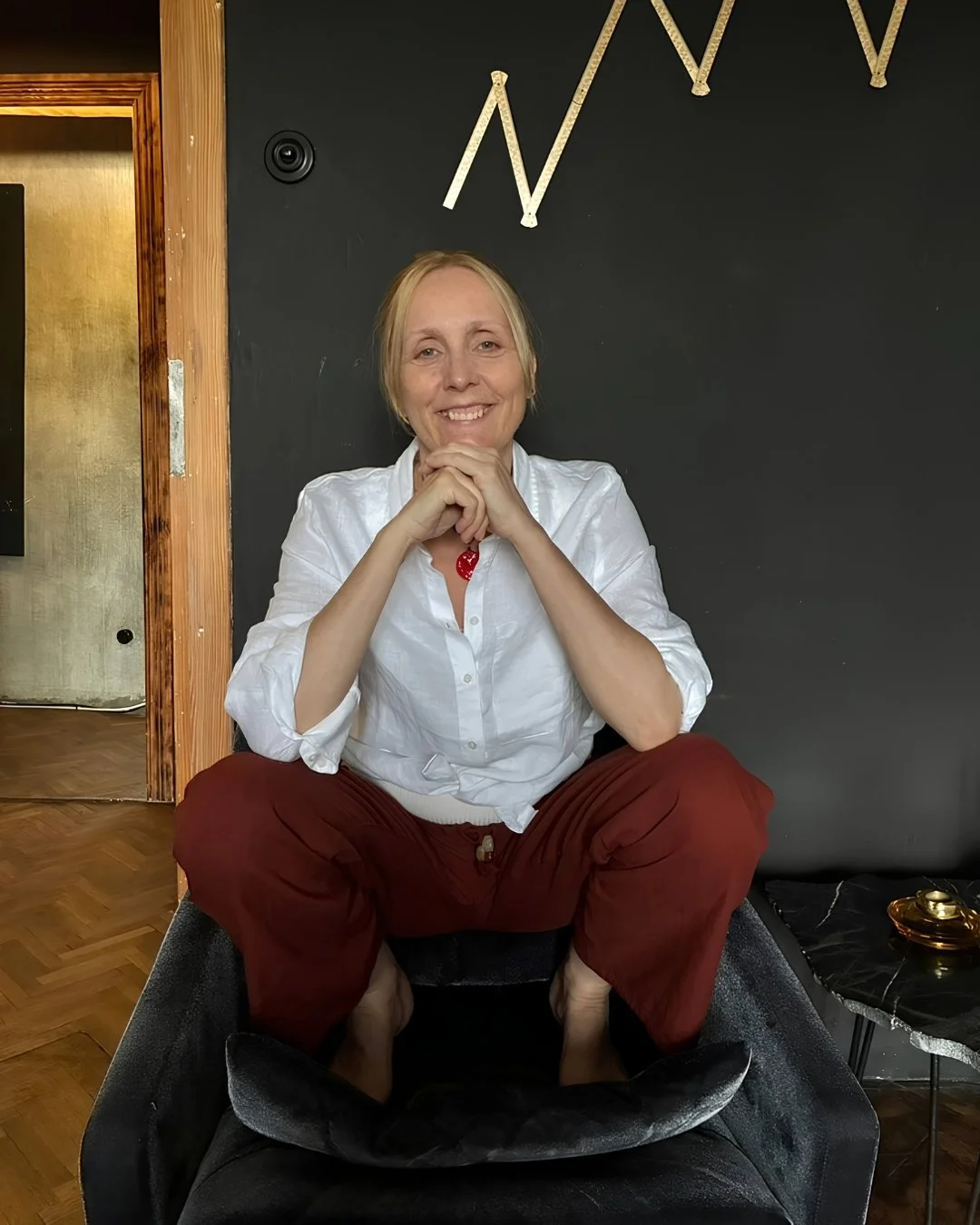 A woman with blonde hair, smiling, sitting cross-legged on a black chair with her hands clasped under her chin, wearing a white shirt and red pants, in a room with wooden flooring and dark walls decorated with gold-colored abstract art.