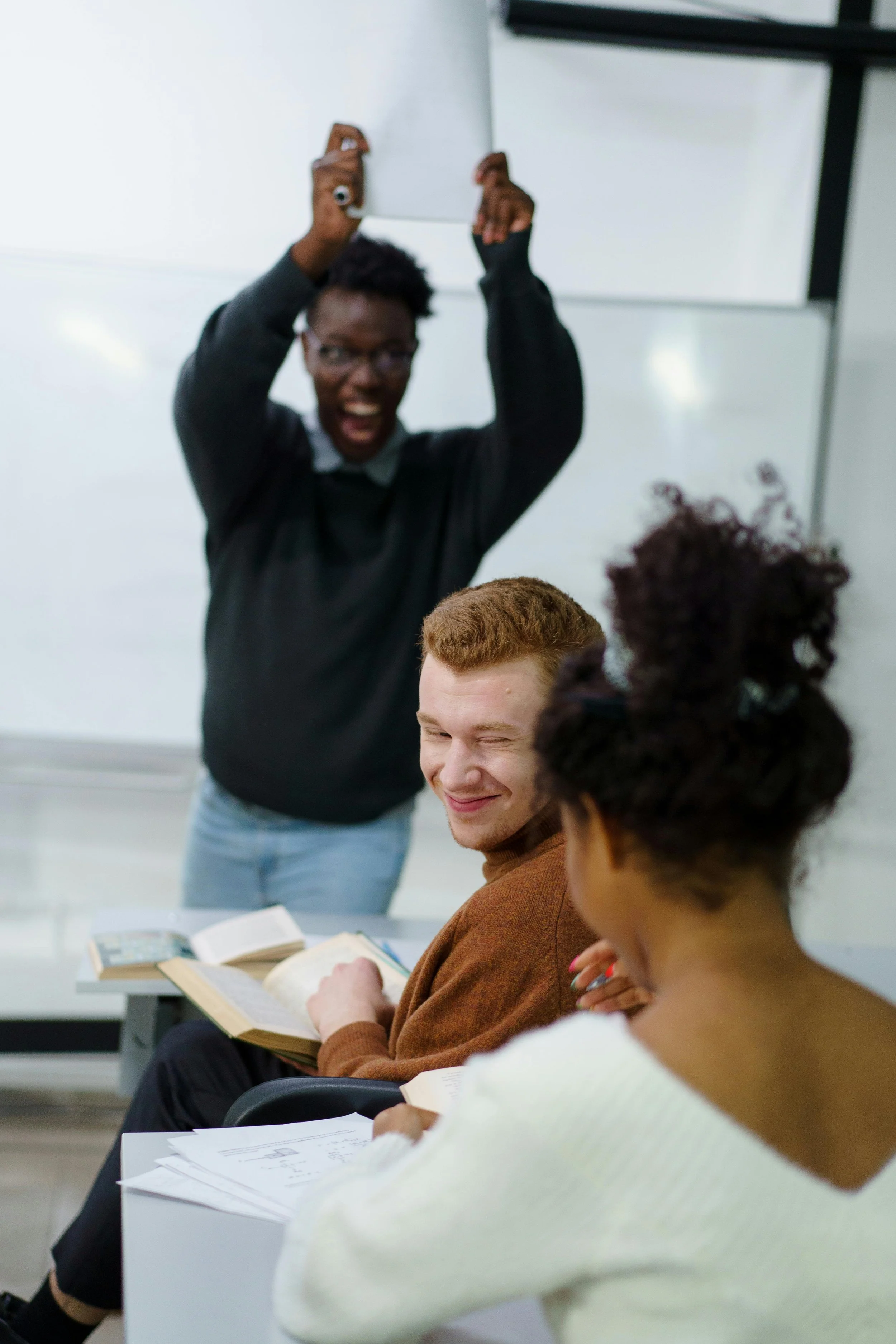 A diverse group of young people in a classroom engaged in a study session. Some are reading, while a woman with short curly hair smiles and holds a book, and a man with glasses and short curly hair is smiling as he reads. A person in the background is standing and raising their arms excitedly.
