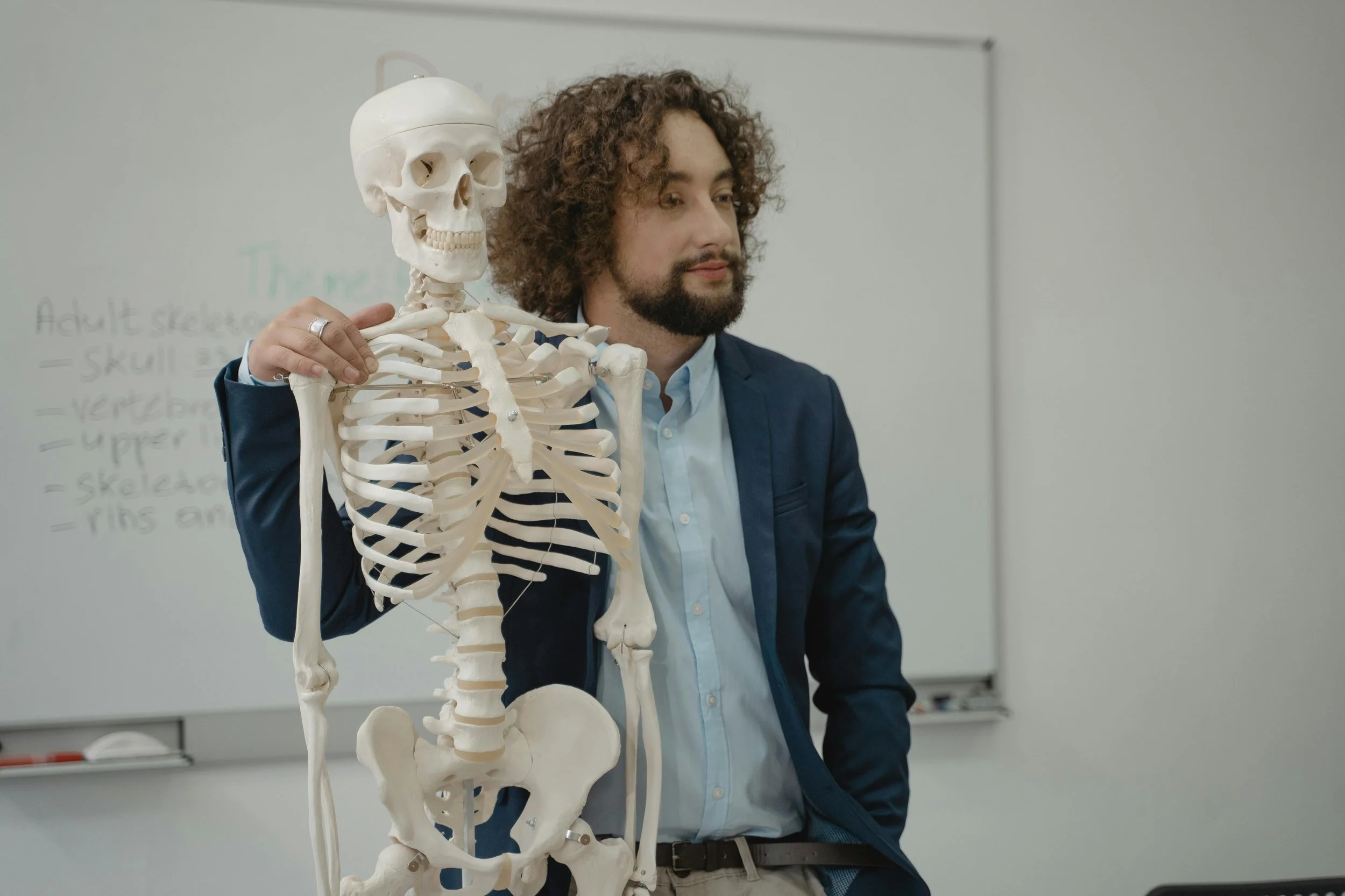 A man with curly hair and a beard in a blue blazer holding a human skeleton model in a classroom setting.