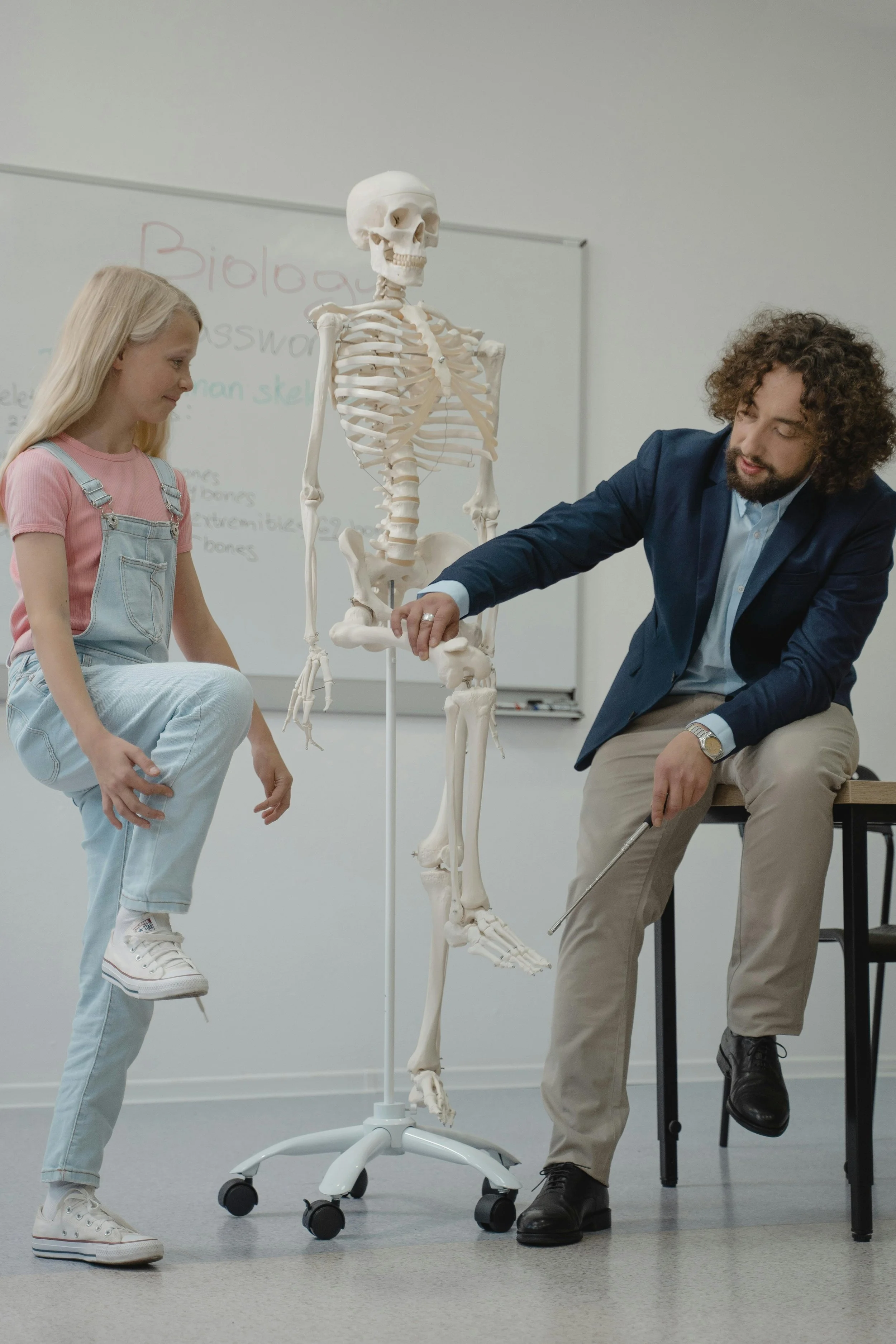 A young girl sitting on a white table with her foot raised, a skeleton model standing next to her, and a man in a suit sitting on a chair pointing at the skeleton. They are in a classroom with a whiteboard in the background.