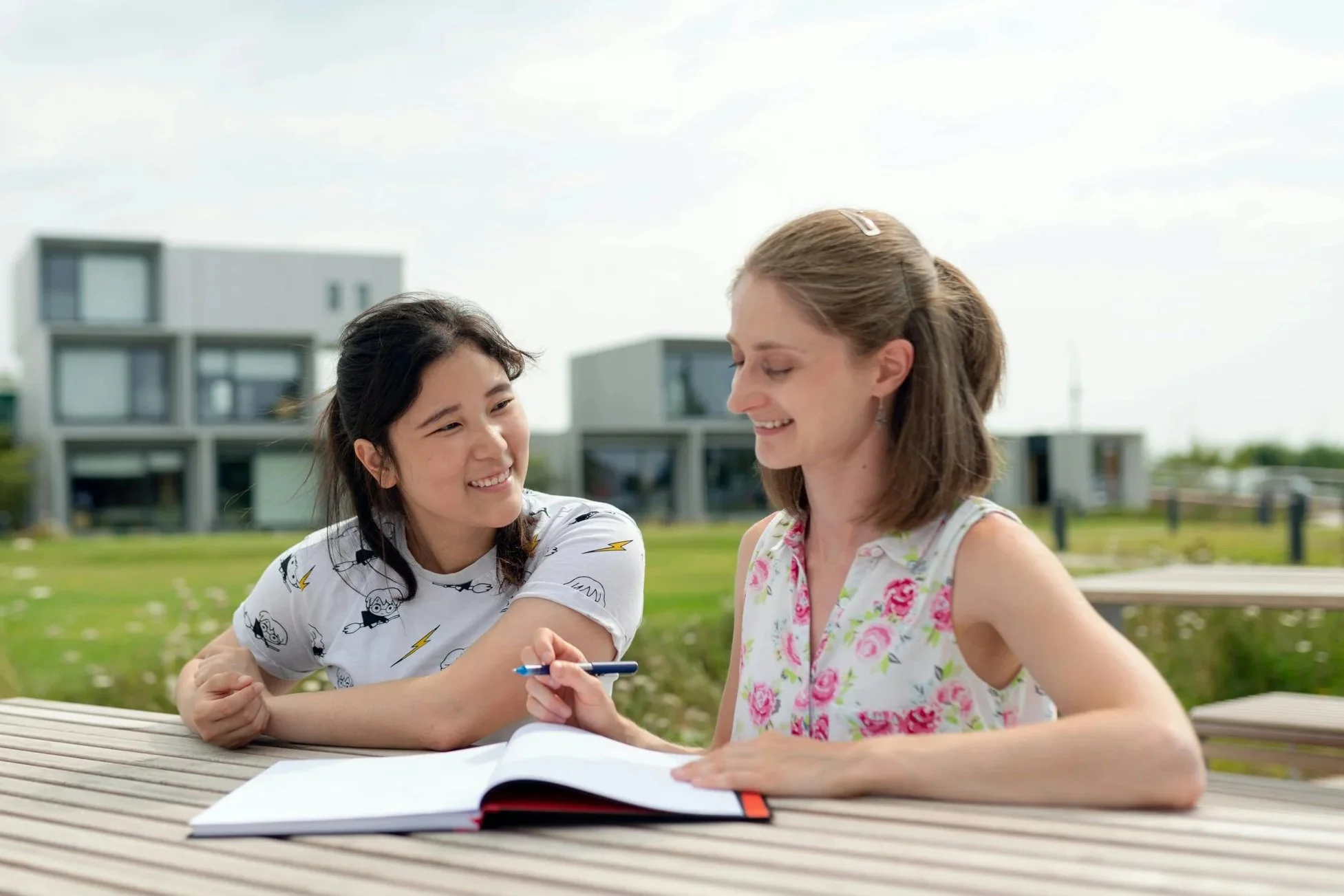 Two women sitting at a picnic table outside, arm wrestling and smiling at each other, with a notebook and pen on the table, and modern buildings in the background.