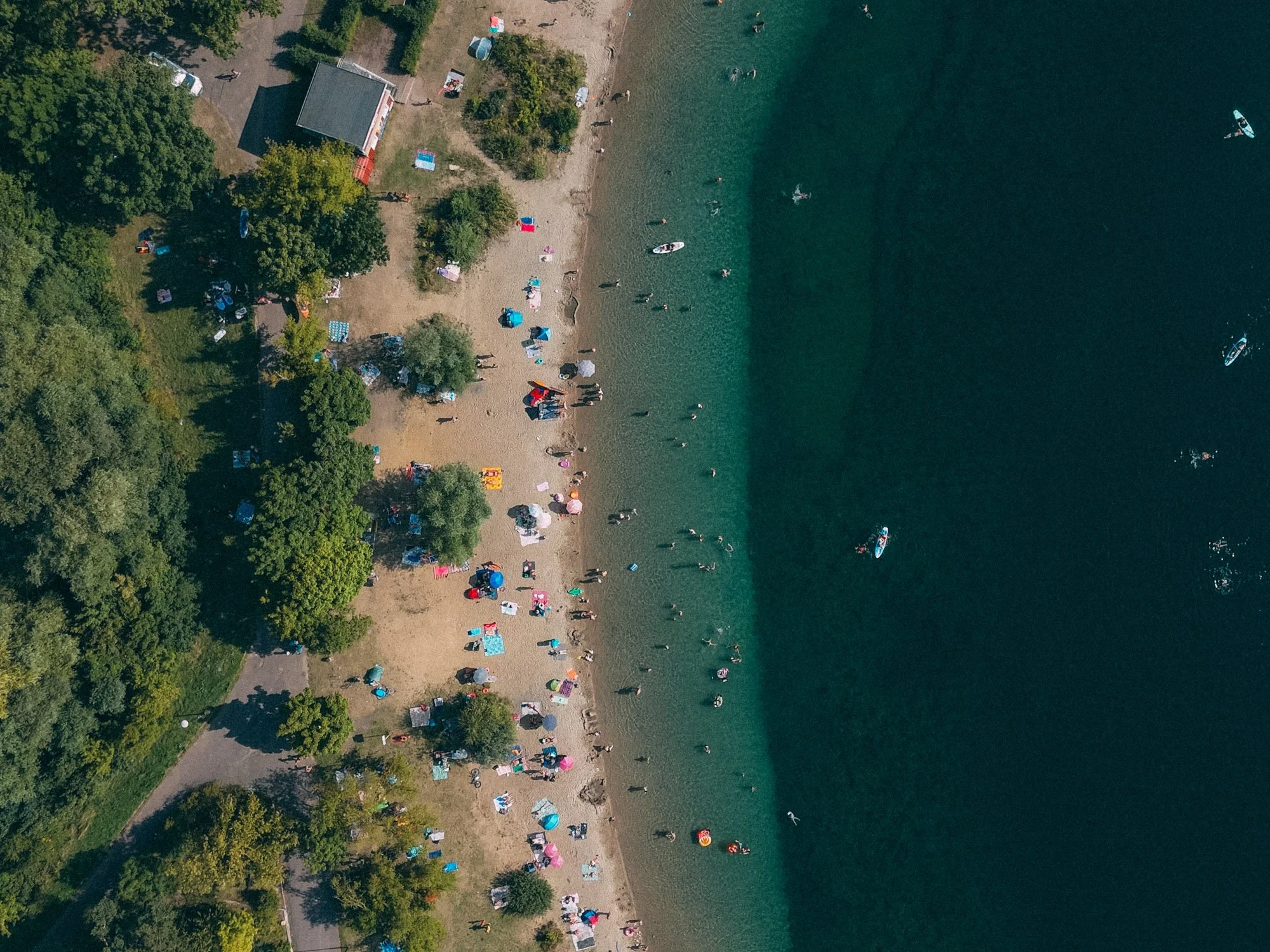Aerial view of a crowded beach with umbrellas, people swimming, and boats on the water next to a wooded shoreline.