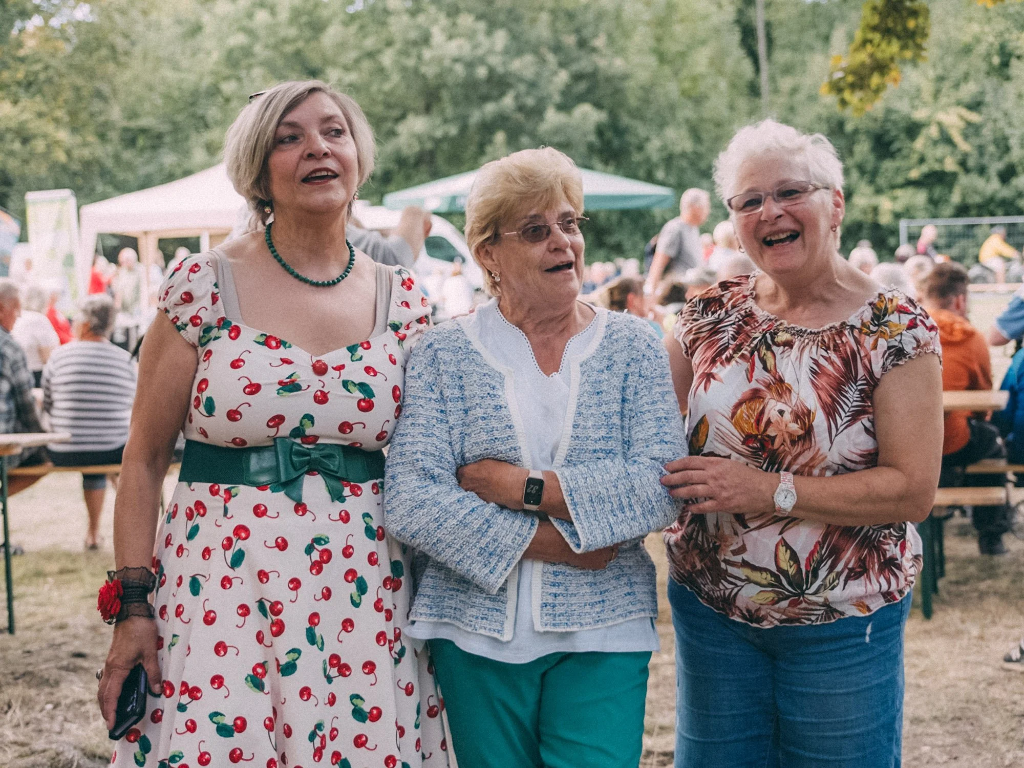 Three elderly women standing outdoors at a gathering, smiling and talking with each other. The background shows trees, tents, and people.