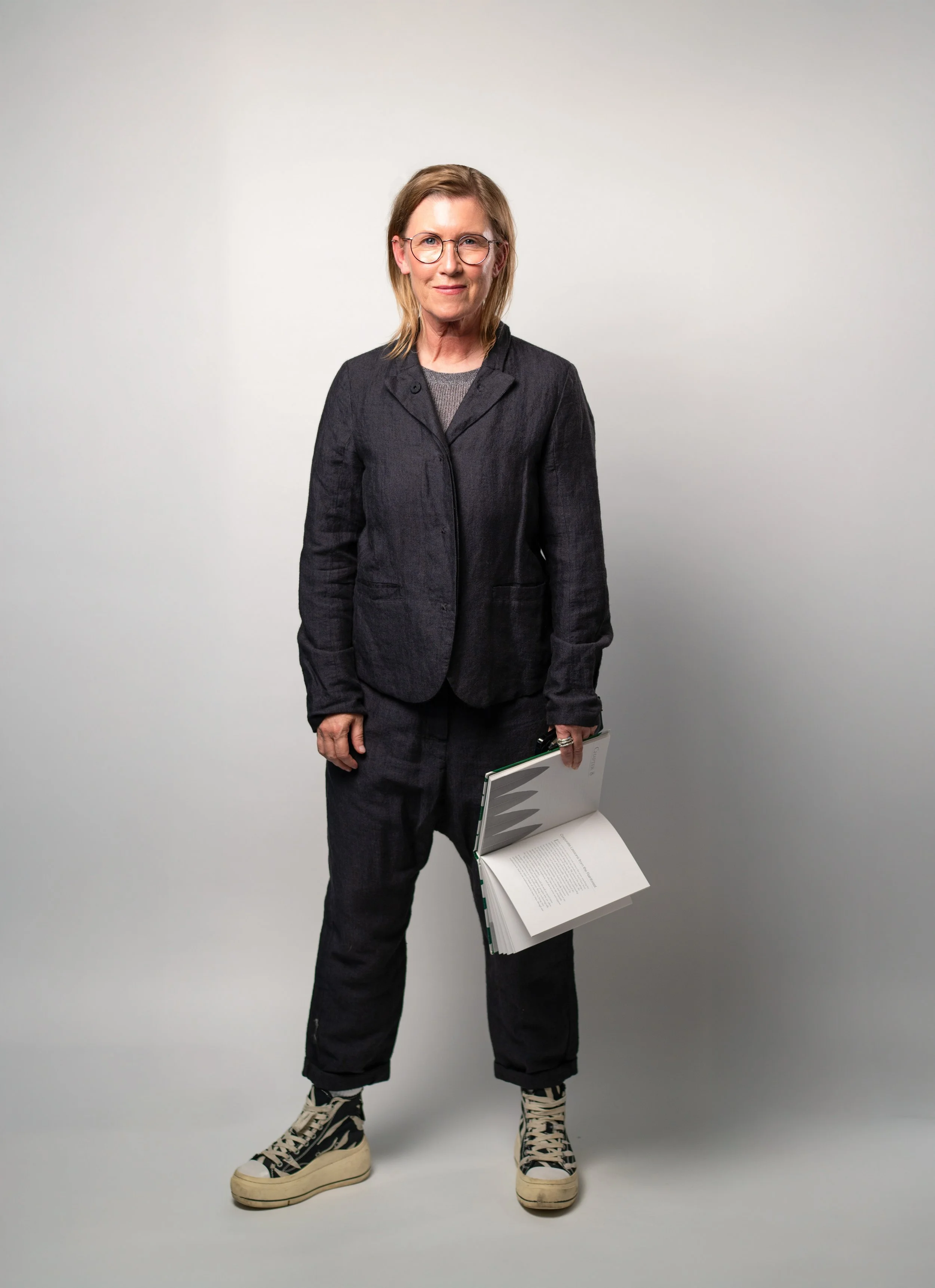 Eine Frau mit Brille trägt schwarze Kleidung, sportliche Schuhe, hält eine offene Buch in der Hand und steht vor weißem Hintergrund.