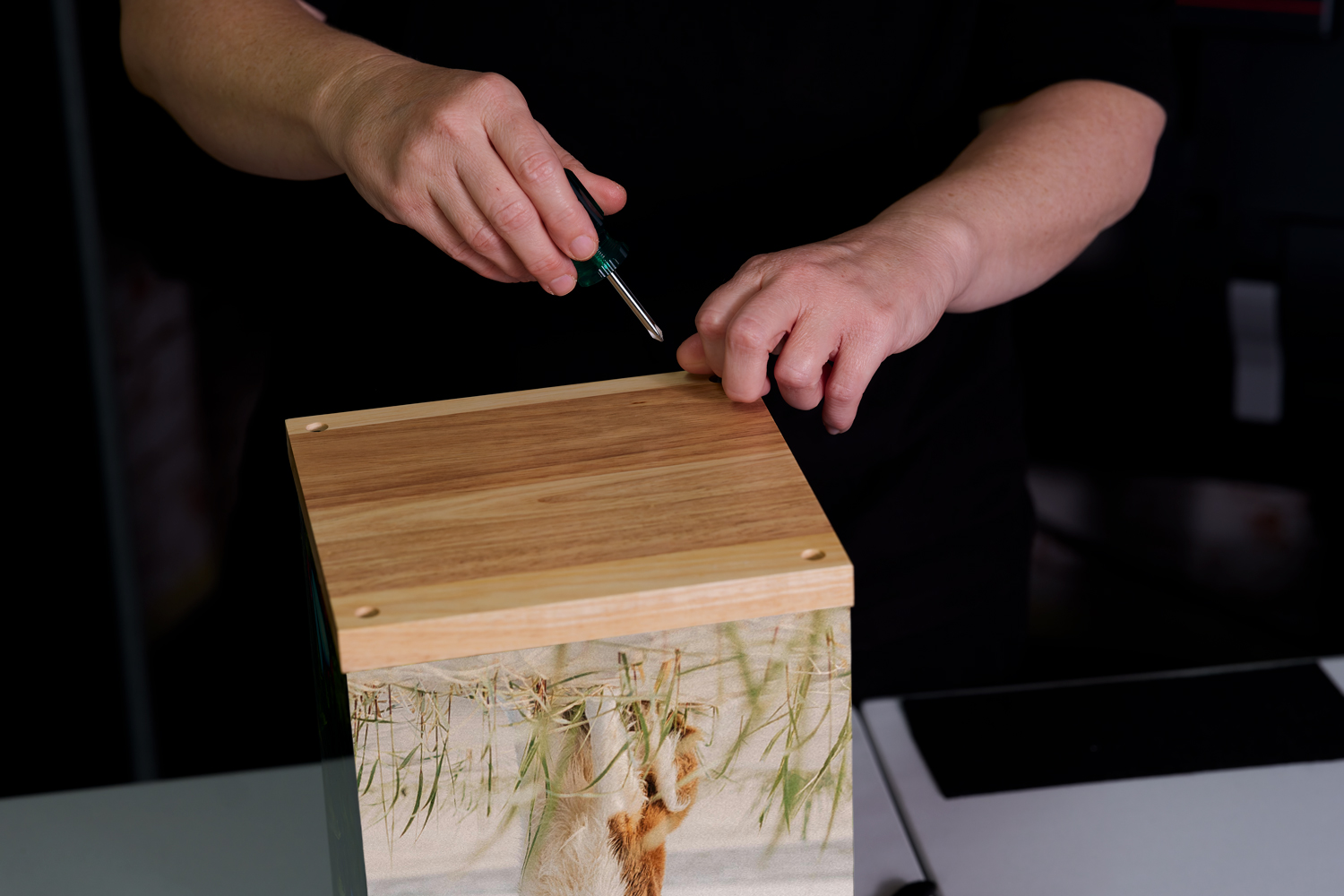 A person is placing a transparent cover on a wooden box that features a nature scene with butterflies and a waterfall.