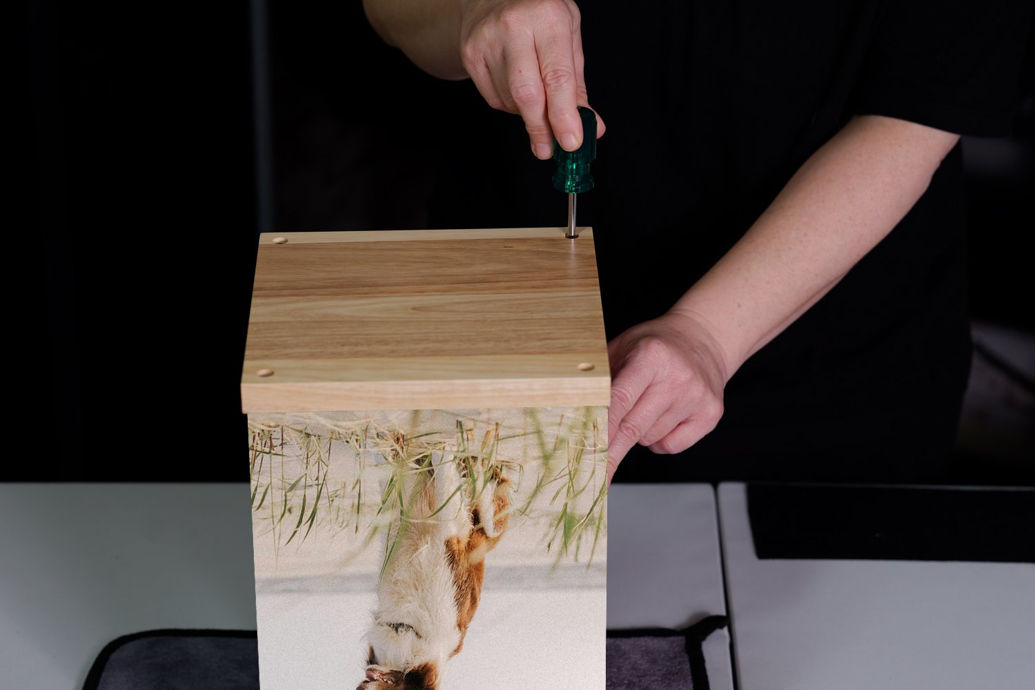 Person using a screwdriver to assemble a wooden box with a nature scene printed on its side, featuring butterflies and a waterfall.