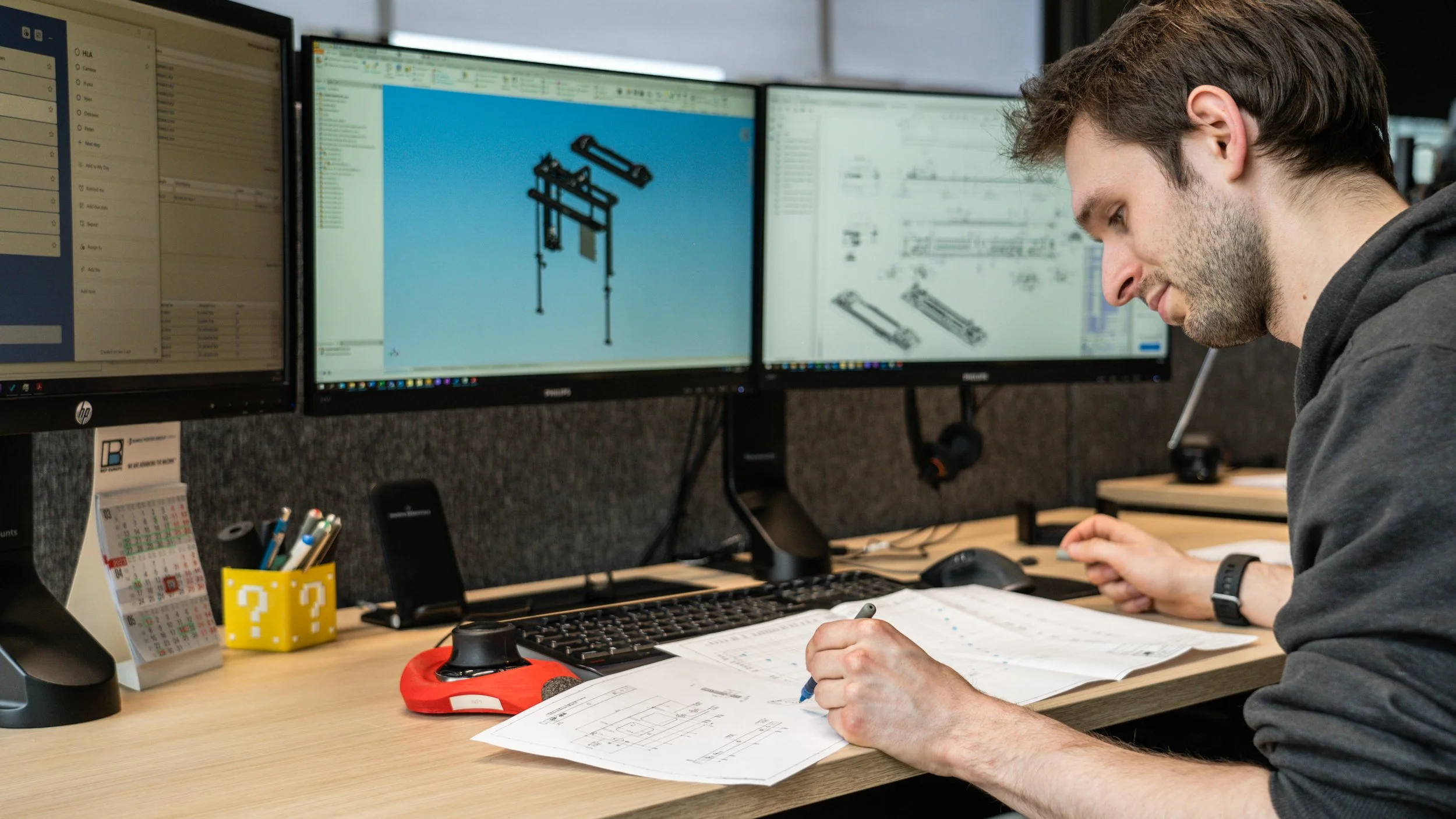 A man working at a desk with three monitors displaying technical drawings and CAD models, surrounded by office supplies.
