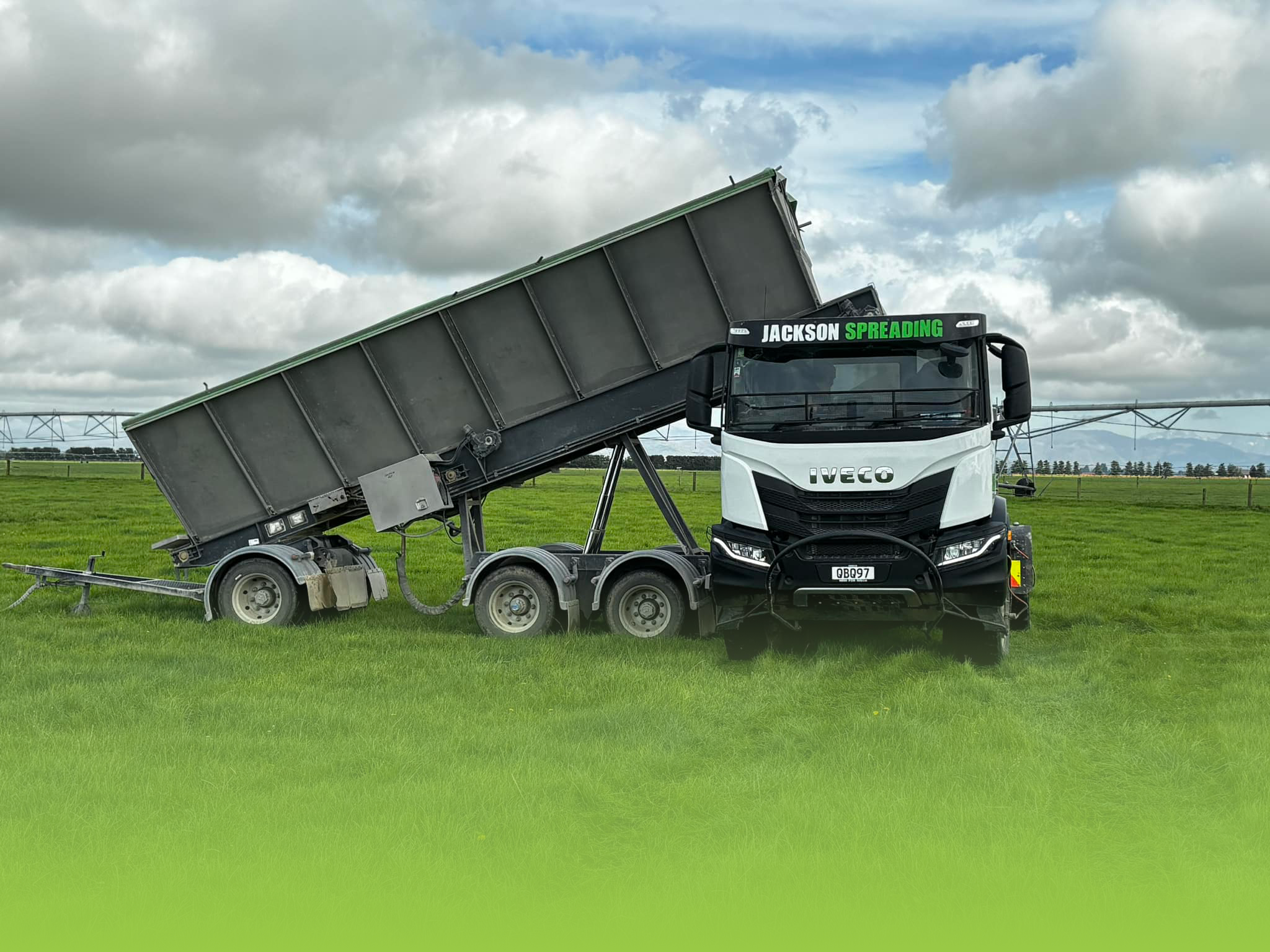 A large white truck with black accents and the name 'IVECO' on the front is seen on a grassy field. The truck's trailer is raised at an angle, revealing a grey underside. The sky is cloudy with patches of blue, and there are structures and trees in the background.
