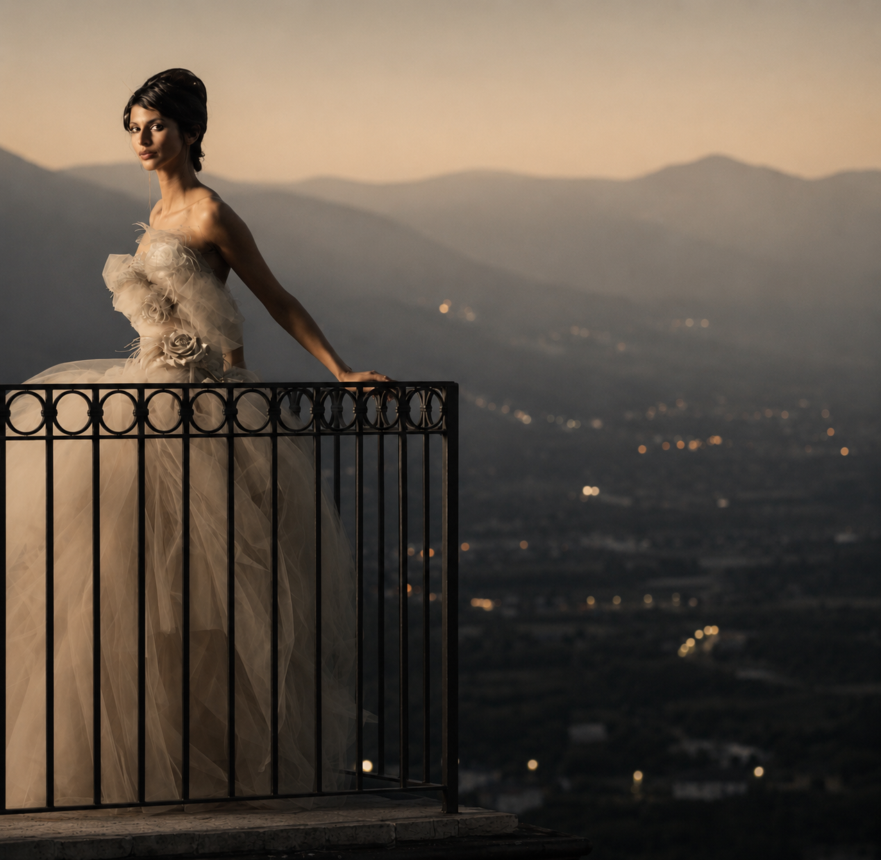 A woman in an elegant beige gown with floral details stands on a balcony overlooking a city and distant mountains at sunset.