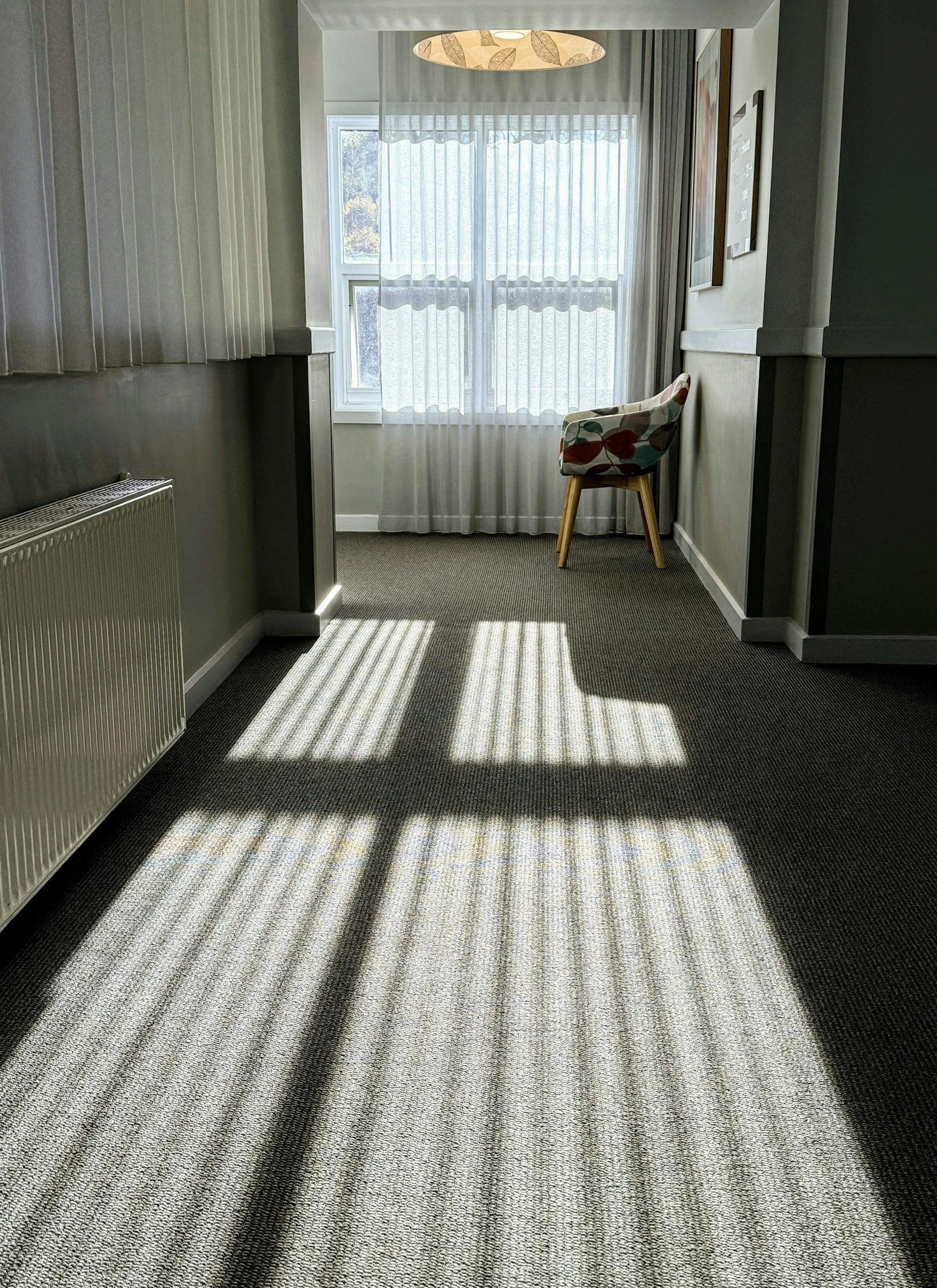Sunlight streaming through a window with white sheer curtains, casting striped shadows on a gray carpeted floor in a corner of a room with gray and white walls and a colorful floral chair.