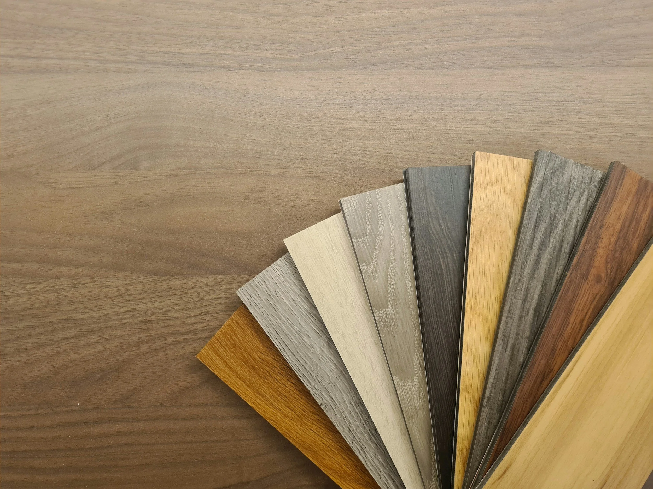 Sample of wood laminate flooring in various shades of brown, gray, and beige, fanned out on a wooden surface.