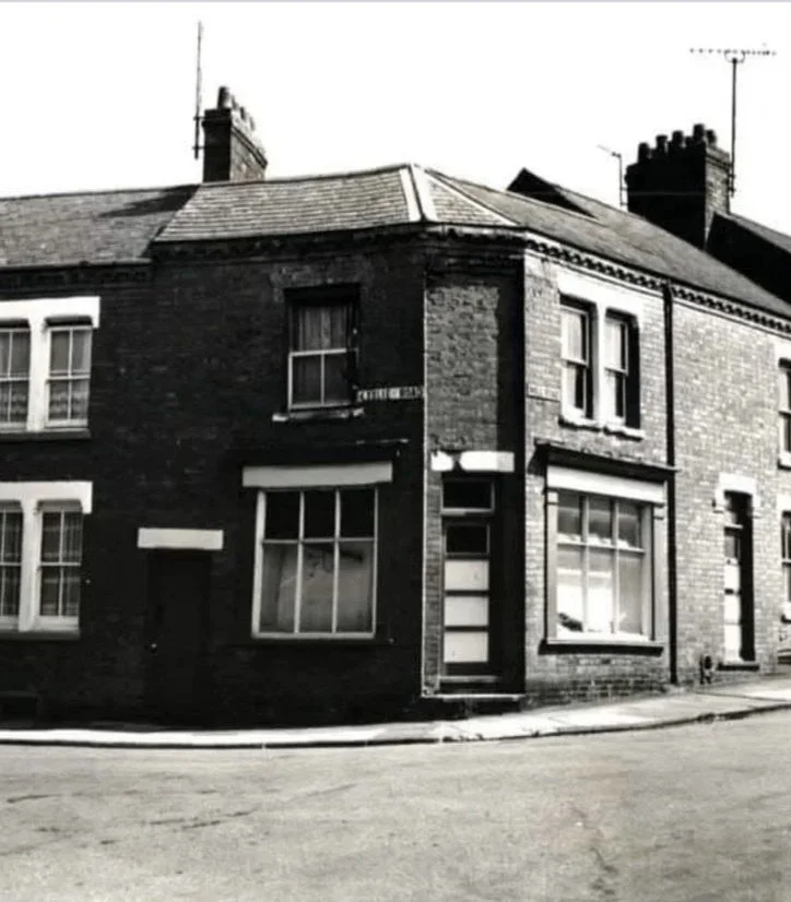 A black and white photo of a corner brick house with large front windows and a small step leading to the door, located on a street corner.