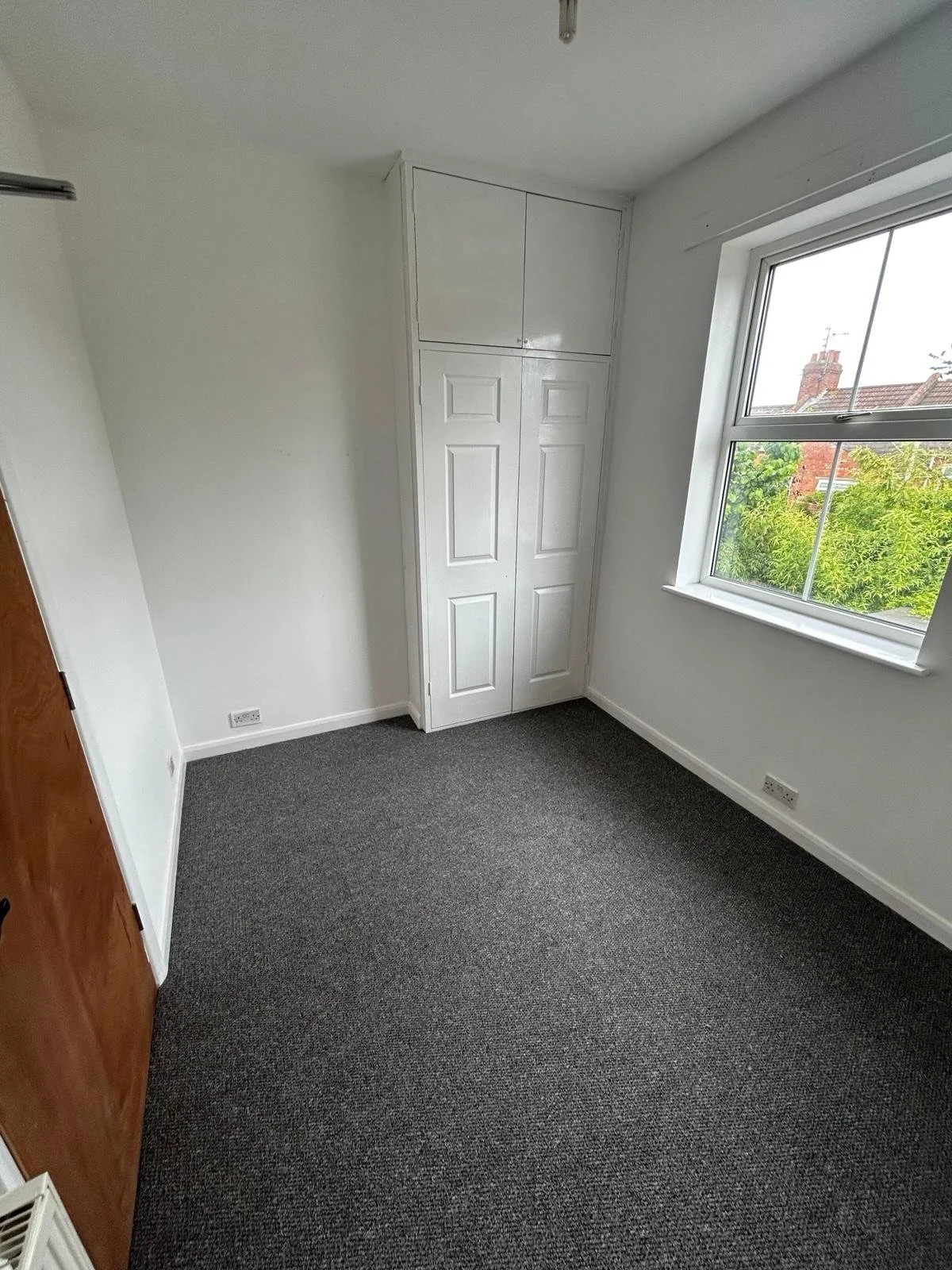 Empty small room with a large window showing trees outside, a white corner wardrobe, a wooden door, and gray carpet flooring.