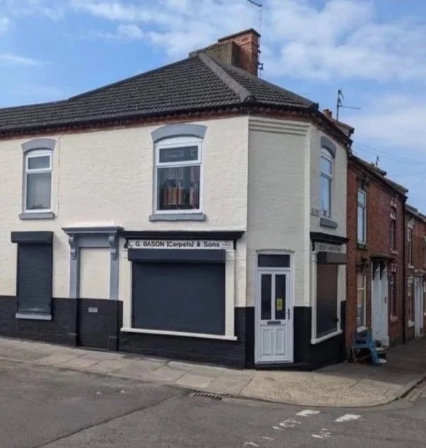 Corner building with white upper walls and black lower walls, featuring three windows with shutters and a door on the right, with a sign that reads "E.J. Bason Carpets & Sons" above a closed shutter, situated on a street under a partly cloudy sky.