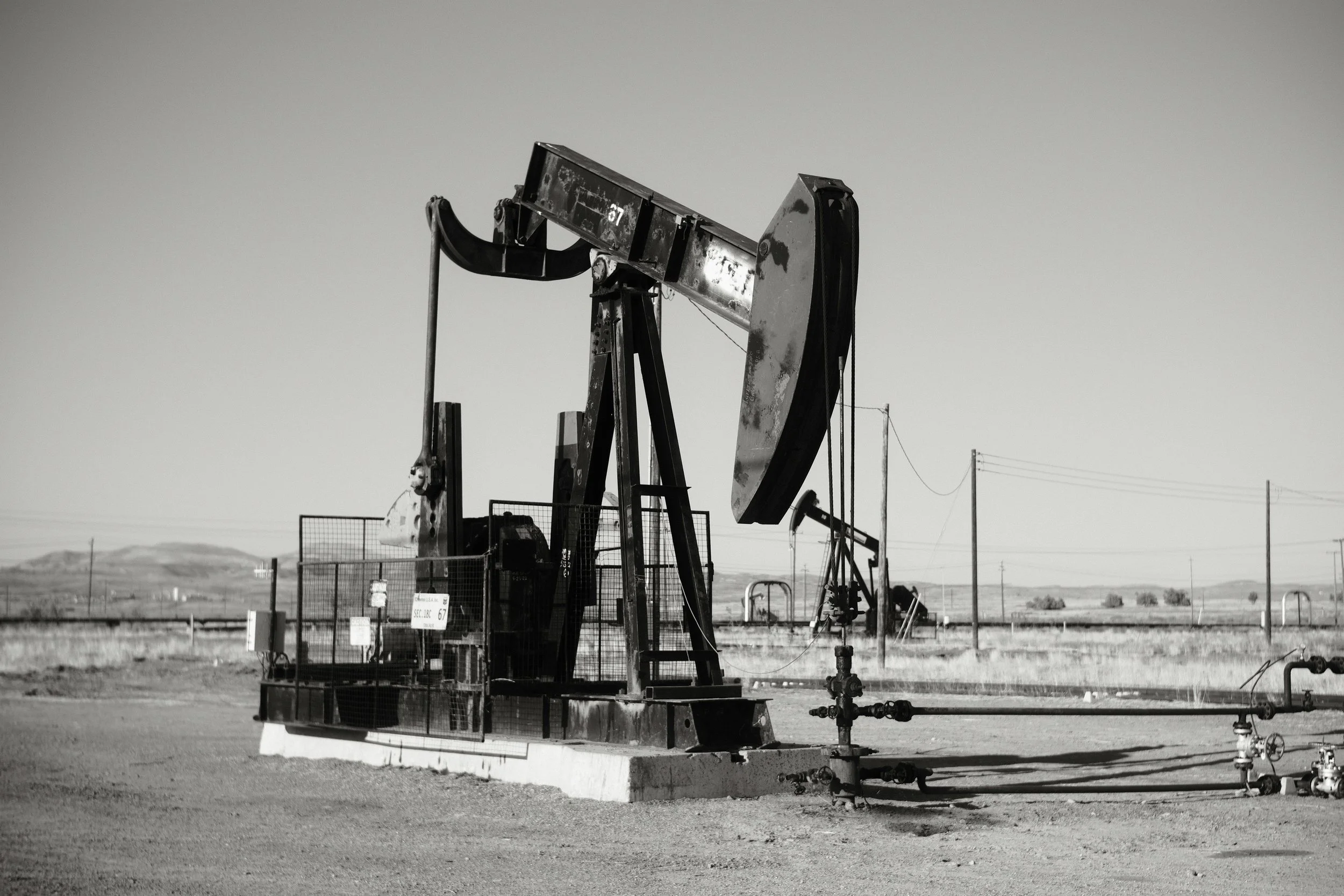Black and white photograph of oil pumpjacks in a flat, open landscape with distant hills and power lines.