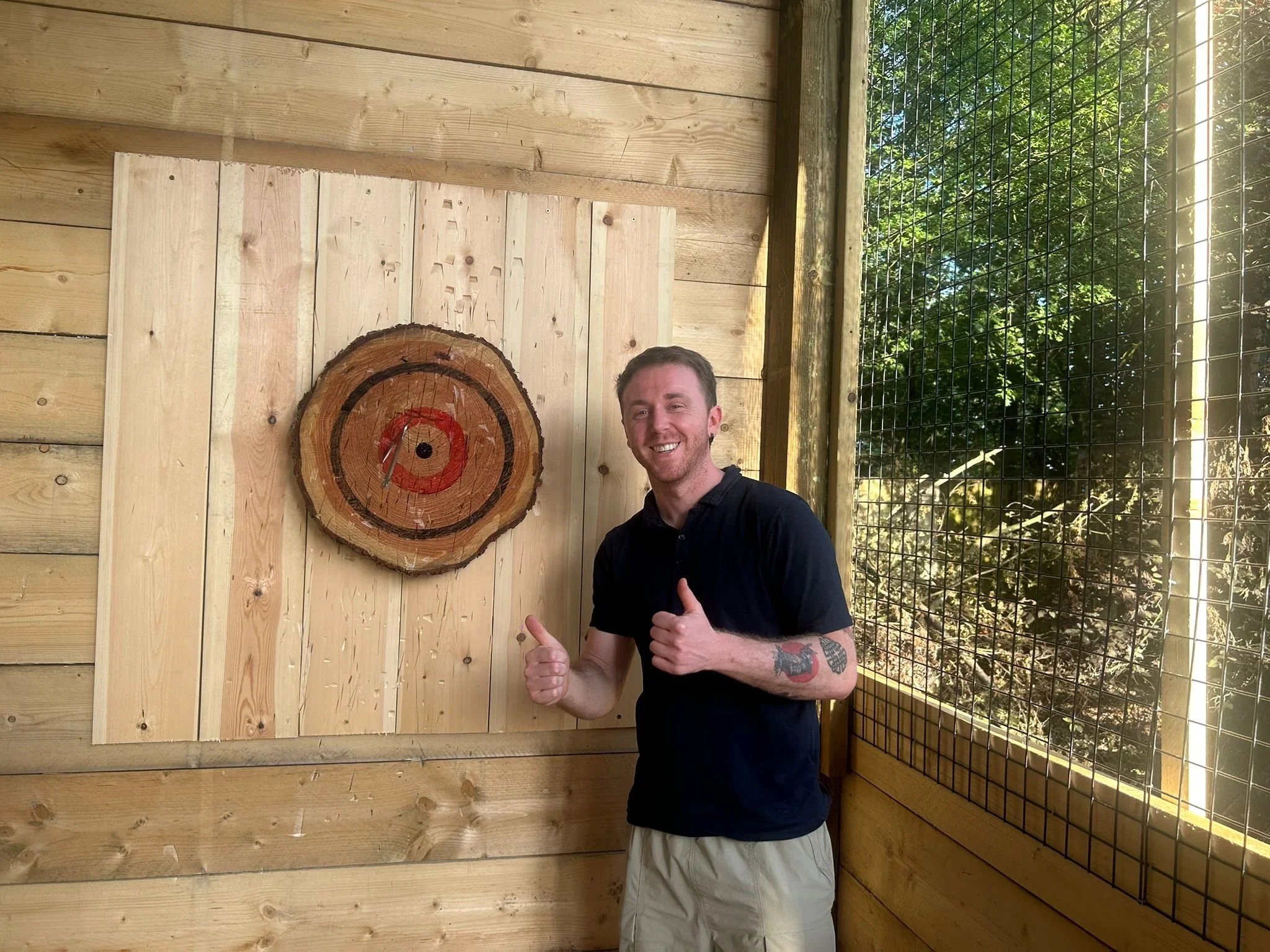 A smiling man with a tattoo on his arm is giving a thumbs-up in front of a wooden wall with a painted target and a netted fence outdoors, with trees visible in the background.