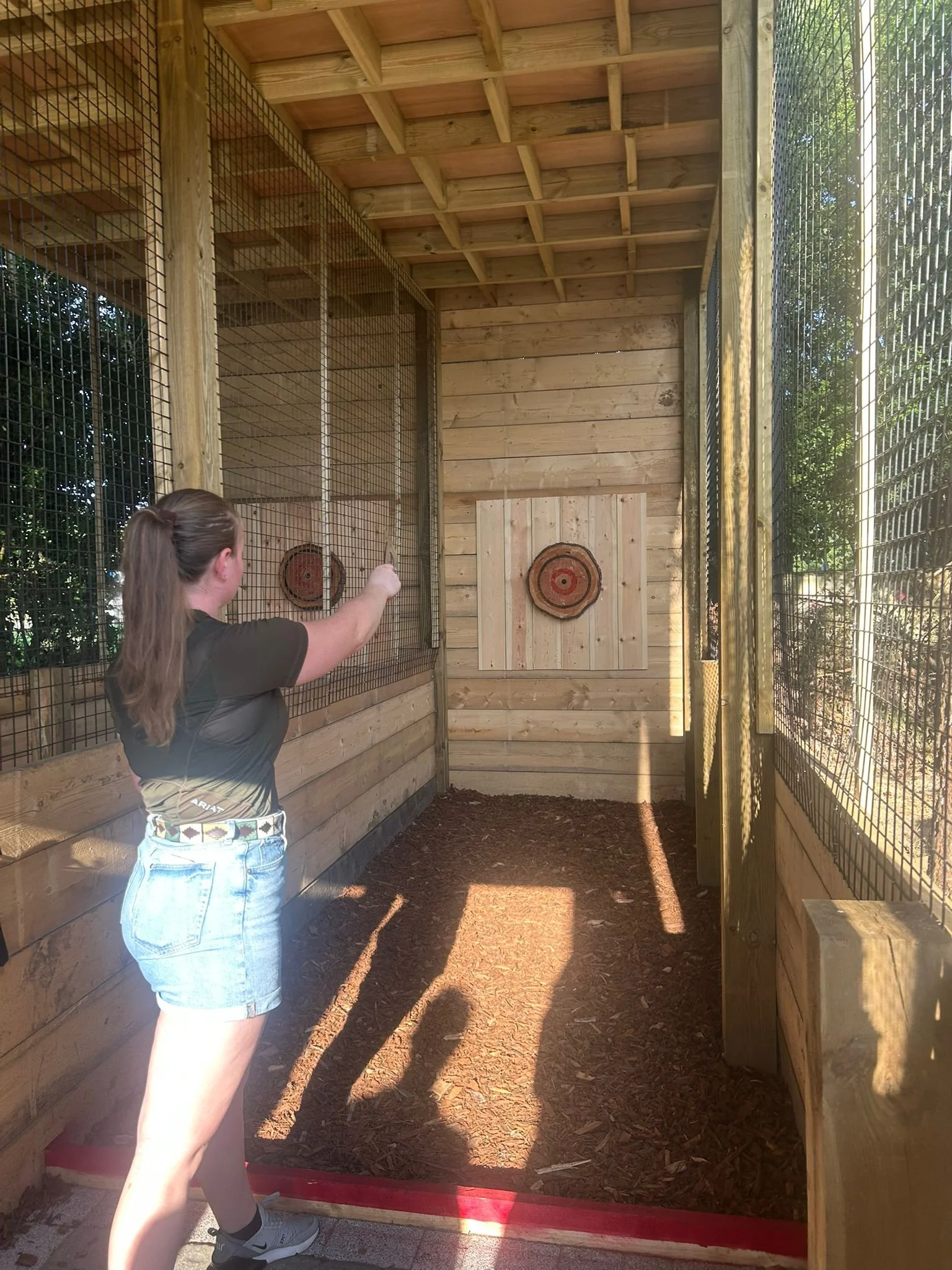 A woman with long brown hair, wearing a black t-shirt, denim shorts, and sneakers, is aiming an arrow at a target inside a wooden and wire mesh enclosed area. The target is mounted on a wooden wall and has concentric red and yellow circles.