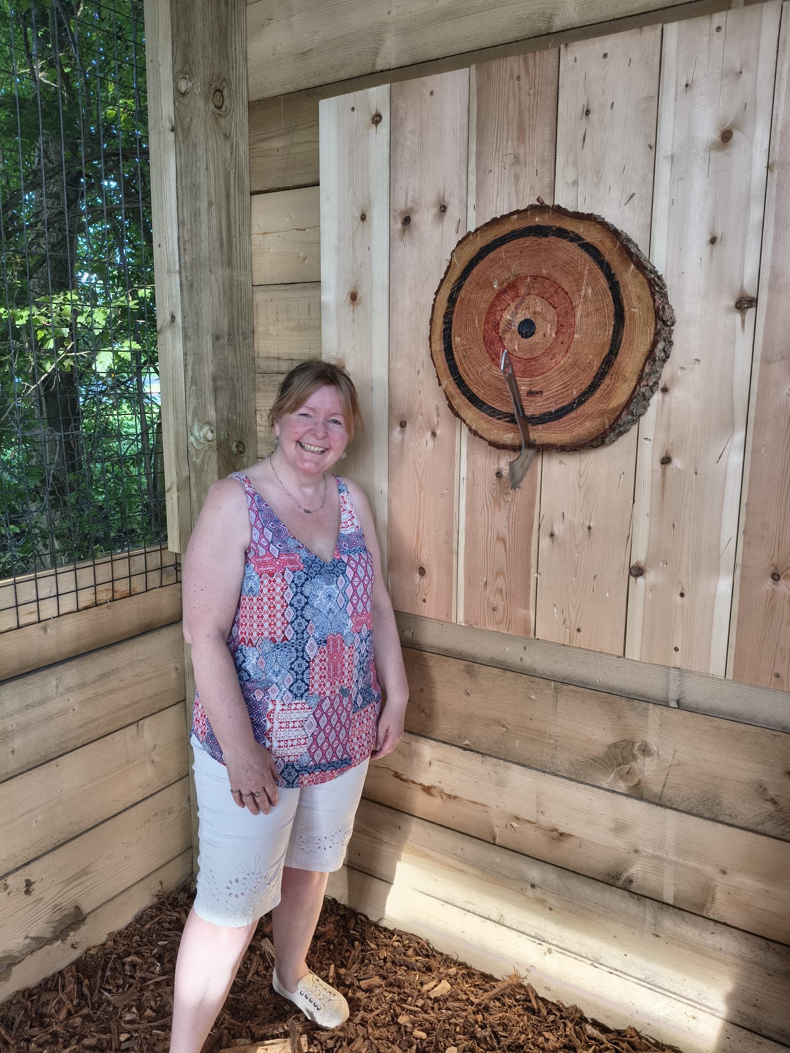 A woman standing in front of a wooden target with a hit bulls eye, inside an outdoor covered. She is smiling and having a great time at Bermuda Blades Axe throwing in Henlow