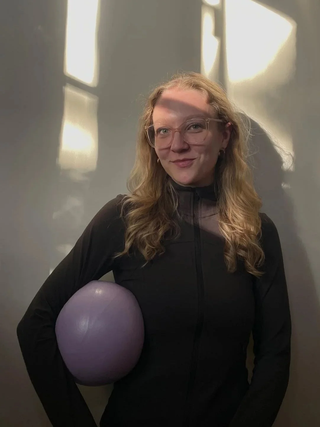 Eine Frau mit langem, gelocktem Haar trägt eine schwarze Jacke und hält einen violetten Ball unter dem Arm. Sie trägt Brille und steht vor einem grauen Hintergrund mit Licht-Reflexionen.