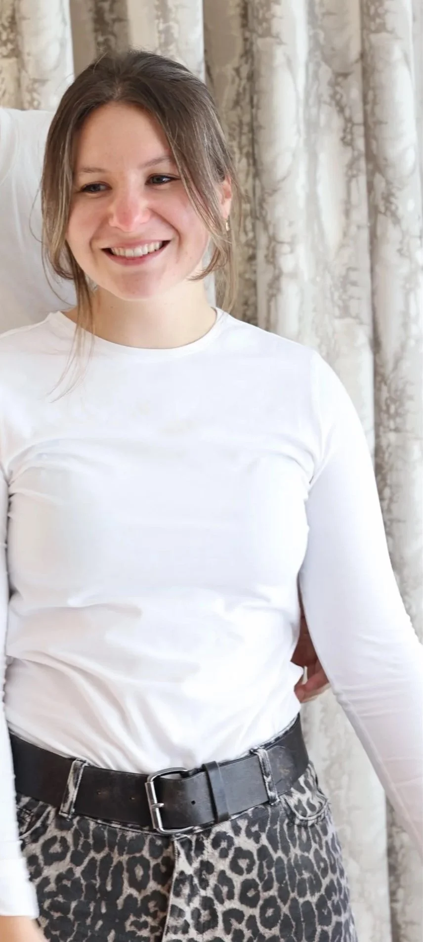 Junge Frau mit braunen Haaren, weiße Langarmbluse, Leopardenmusterhose, Schwarzer Gürtel, stehende Pose vor dekorativen Gardinen.
