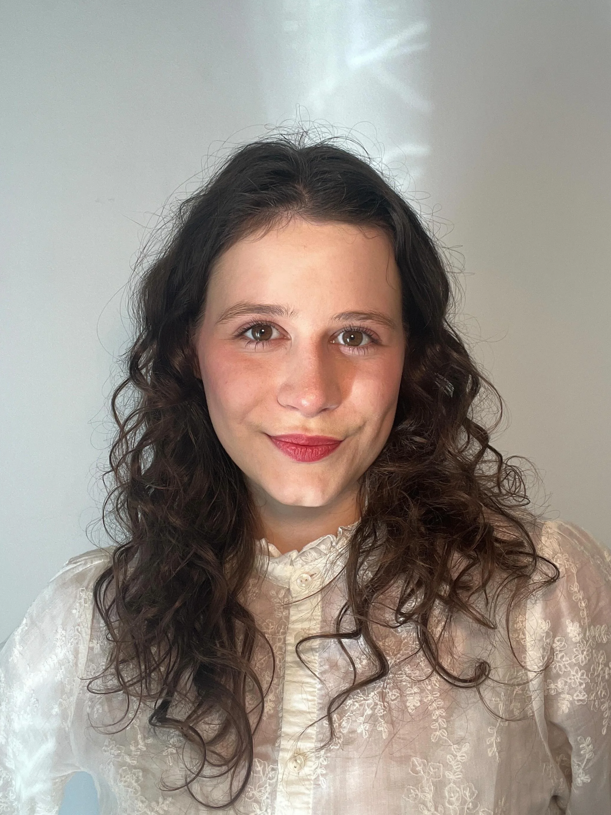 Eine junge Frau mit braunen, lockigen Haaren trägt eine weiße Bluse mit Blumenmuster.