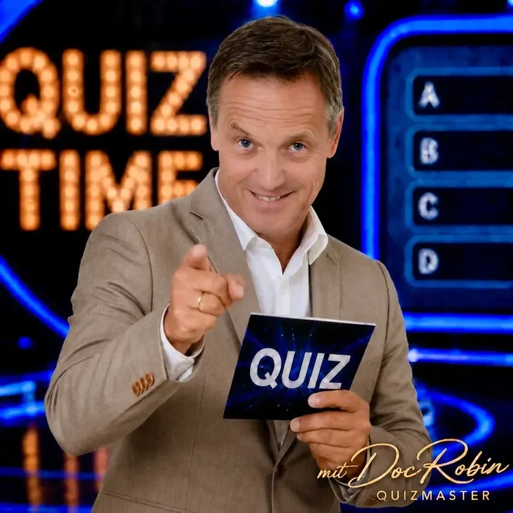 Ein Moderator in einem TV-Studio hält eine Quizkarte mit dem Wort "QUIZ" und lächelt in die Kamera, im Hintergrund ein leuchtendes Spielshow-Set.