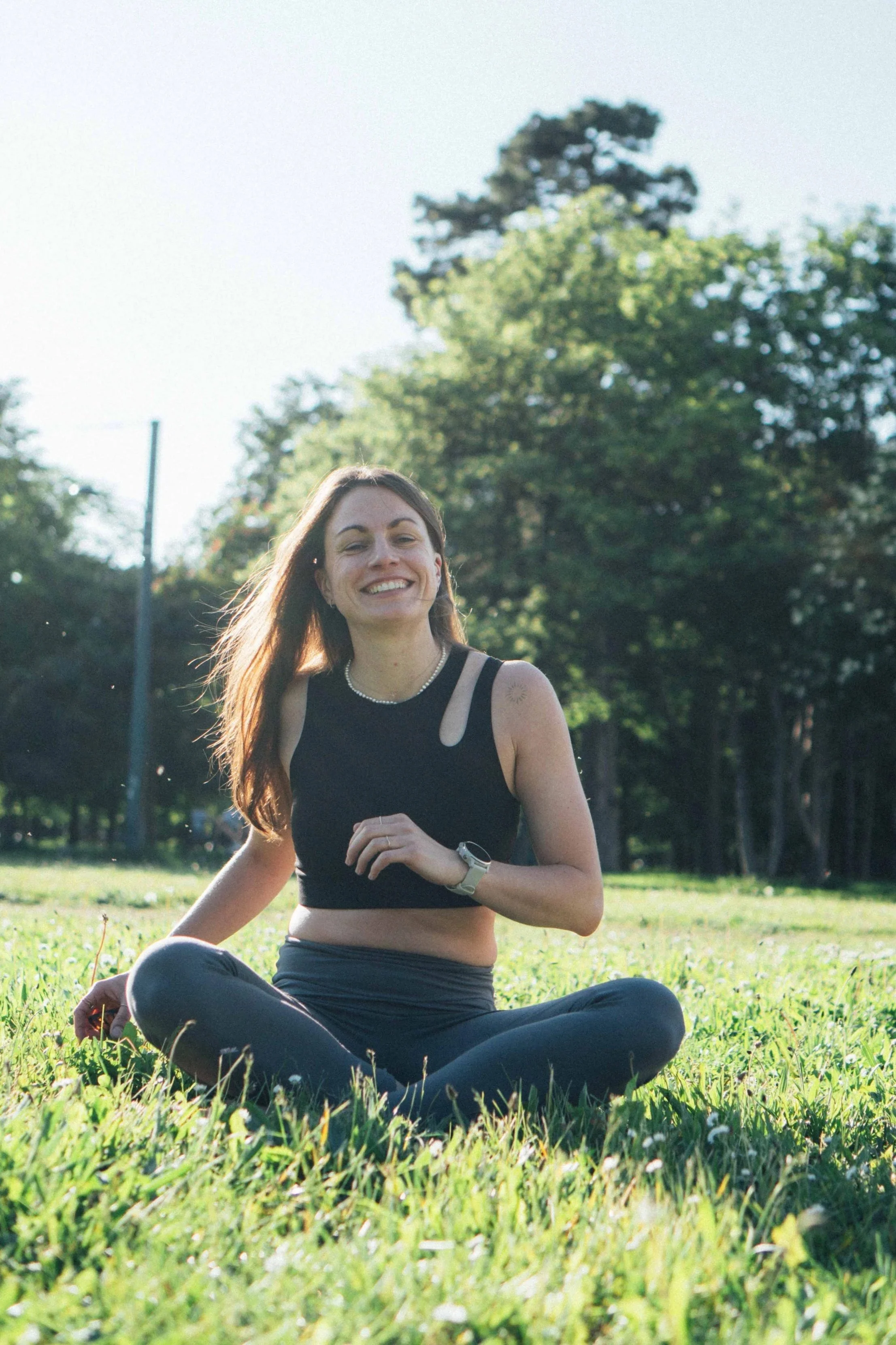 Lächelnde Frau beim Yoga im Park an einem sonnigen Tag, sitzend auf Gras, umgeben von Bäumen.