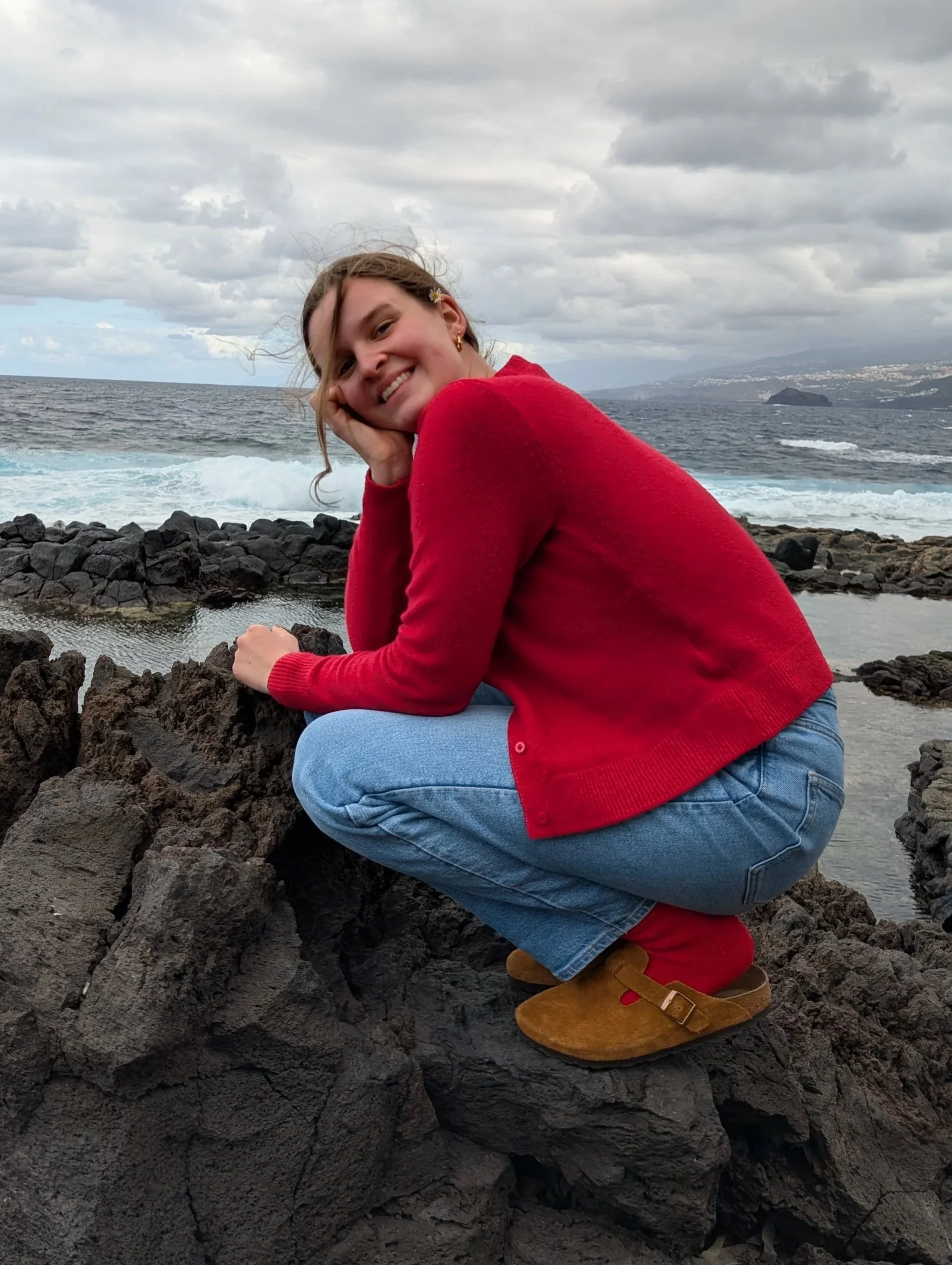 Lächelnde junge Frau in rotem Pullover, blauer Jeans, braunen Schuhen, kniet auf schwarze Lava-Steine am Meer, Blick auf den Horizont, bewölkter Himmel.