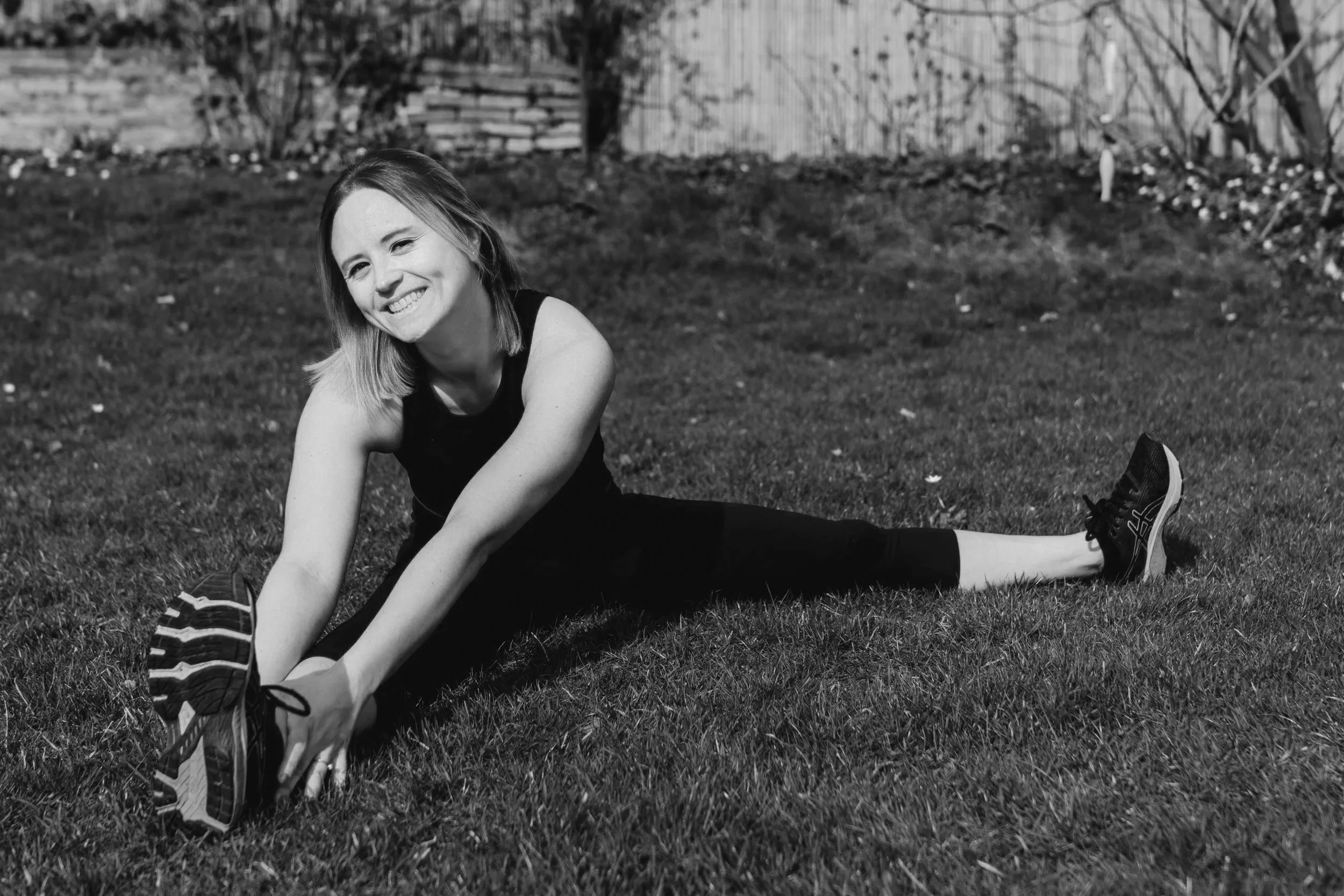 Frau dehnt sich im Freien auf einer Wiese, lächelnd, in sportlicher Kleidung, schwarze und weiße Turnschuhe, im Hintergrund Bäume und eine Holzkonstruktion.