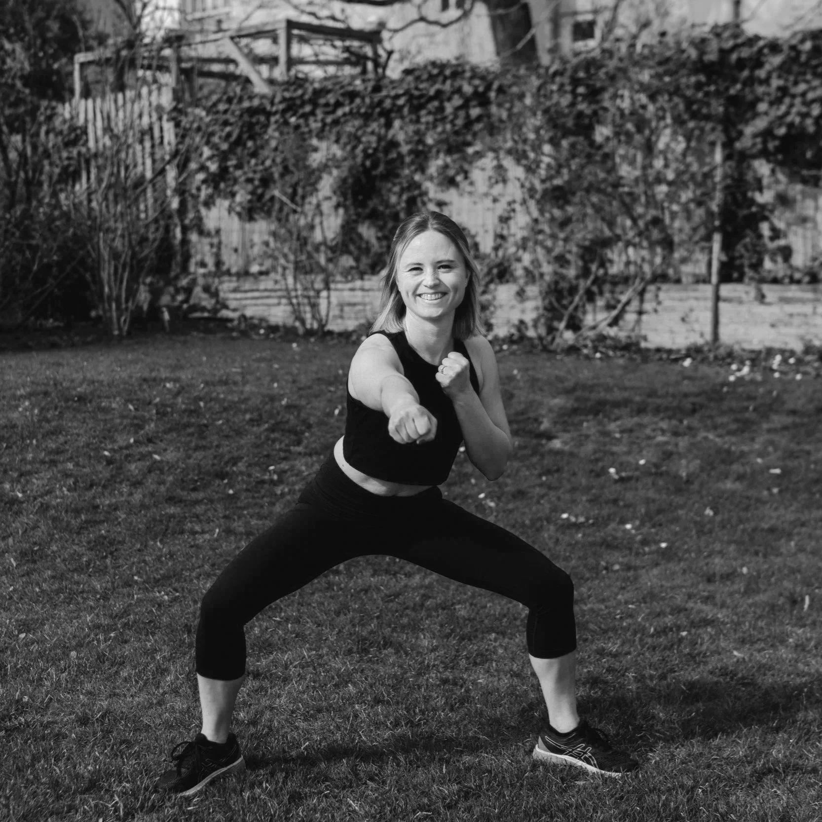 Eine lächelnde junge Frau im Sportoutfit, die eine Kampfhaltung einnimmt, bei einer Trainingseinheit im Freien auf einem Rasen. Im Hintergrund ist eine Gartenmauer mit bewachsenen Pflanzen zu sehen.