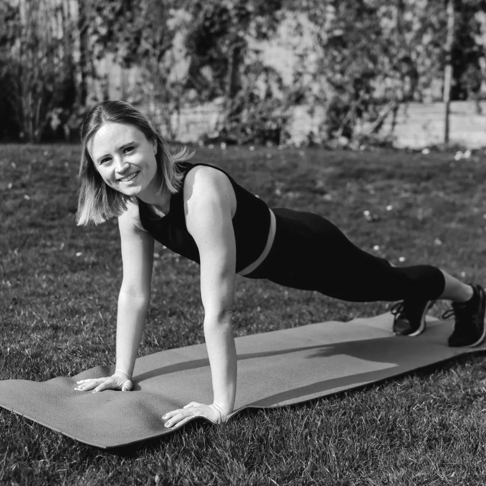 Eine junge Frau macht Yoga im Park auf einer Matte, lächelt und schaut in die Kamera, umgeben von Bäumen und Gräsern.