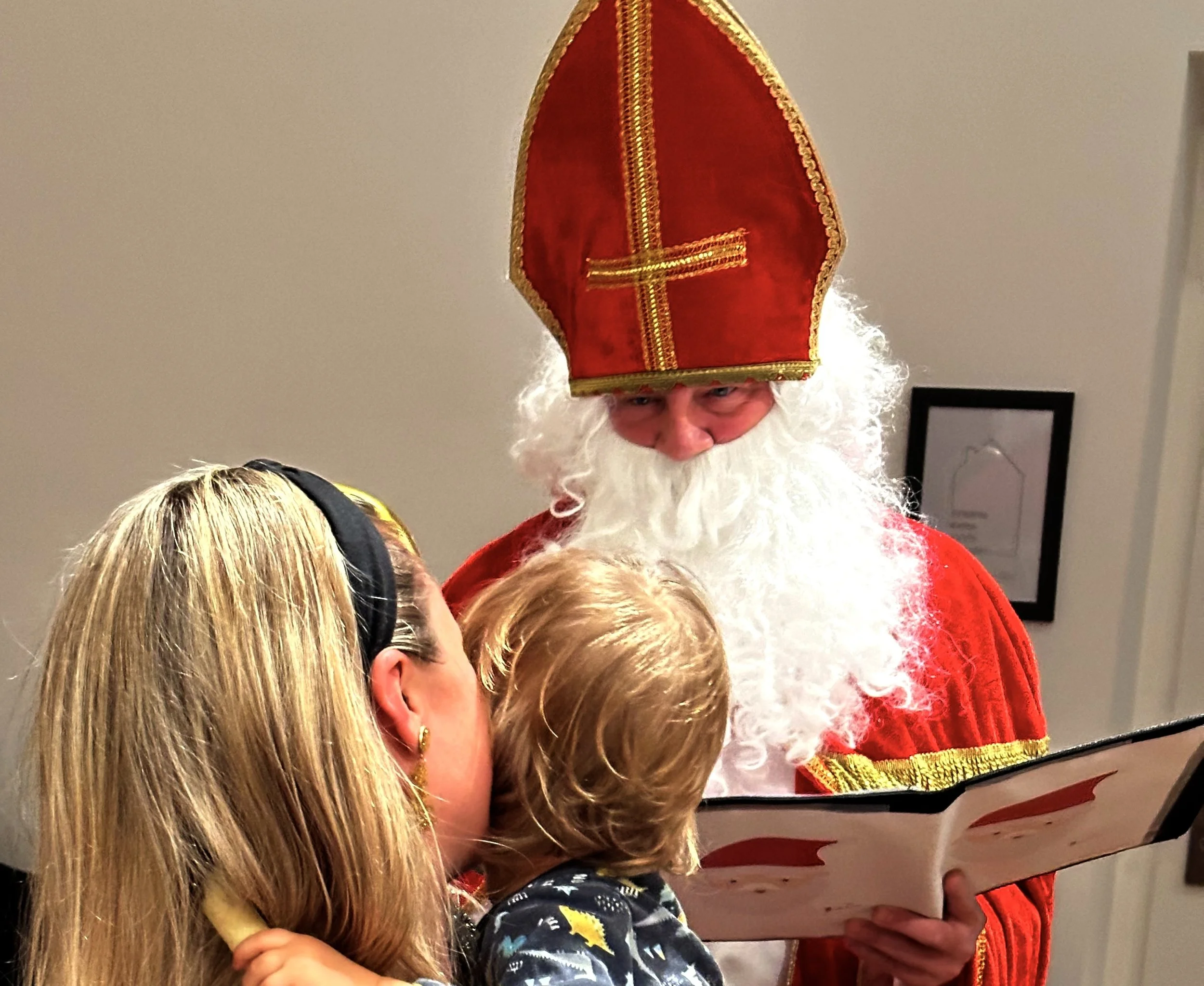 Ein Mann in einem roten Gewand mit goldenen Verzierungen und einem hohen roten Hut mit goldener Spitze, der eine Gruppe von Kindern begrüßt.