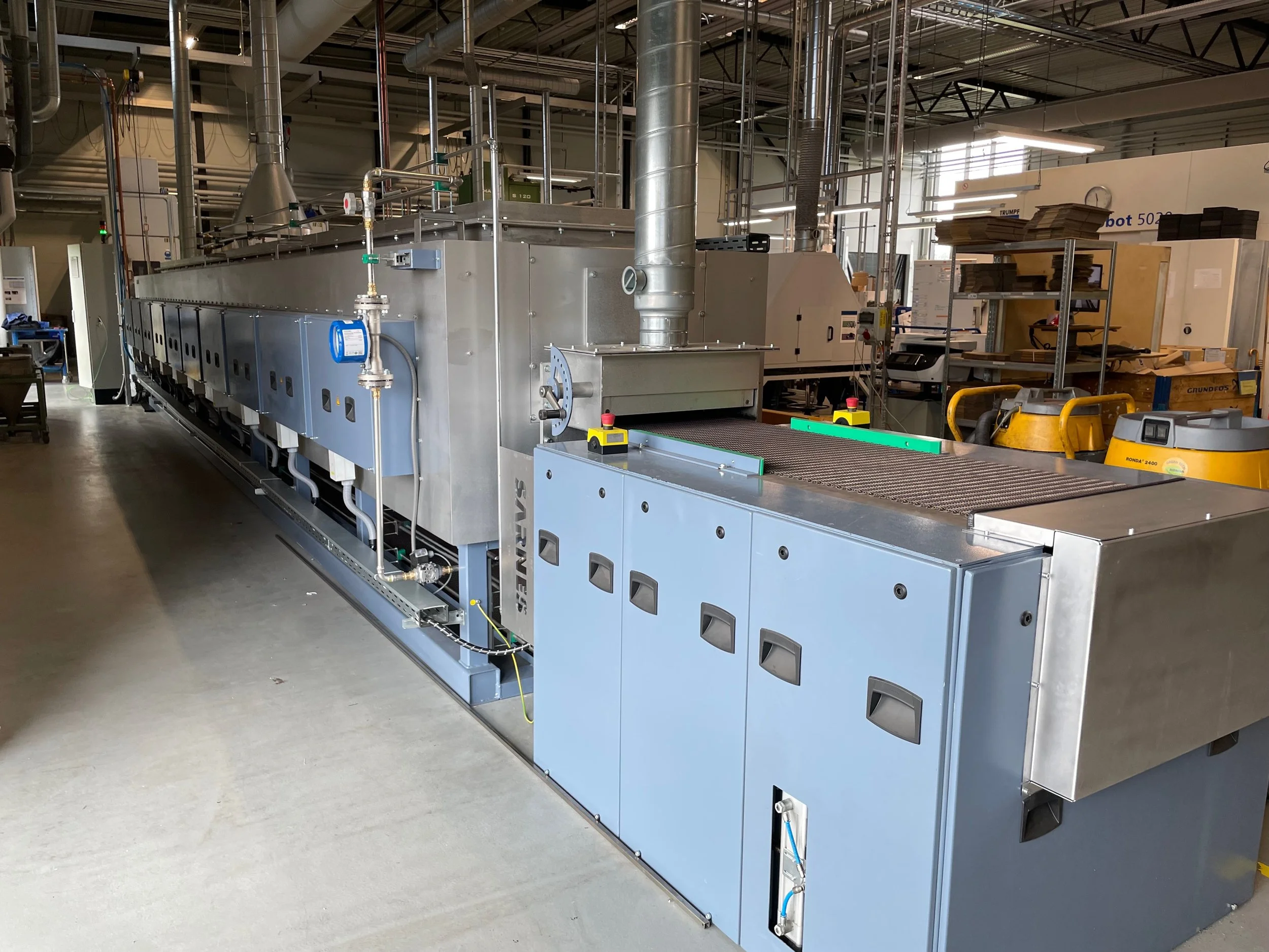 Große industrielle Maschine in einem Werkraum, umgeben von Metallrohren, Kontrollschaltern und Arbeitsmaterialien.