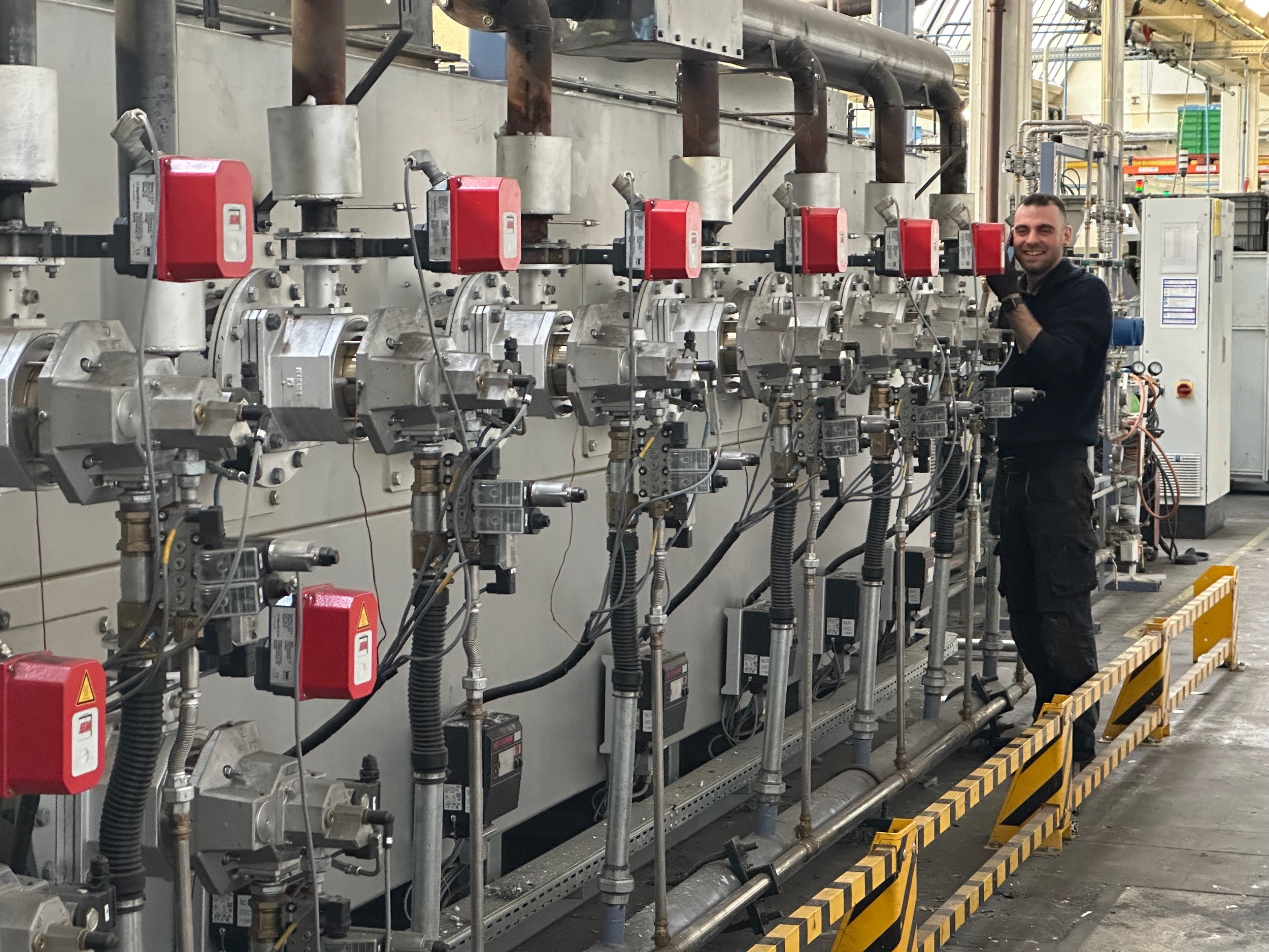 Ein Mann in Arbeitskleidung steht neben einem Sinterbandofen mit mehreren roten Sicherheitsschaltern und Metallrohren in einer Fabrikhalle.