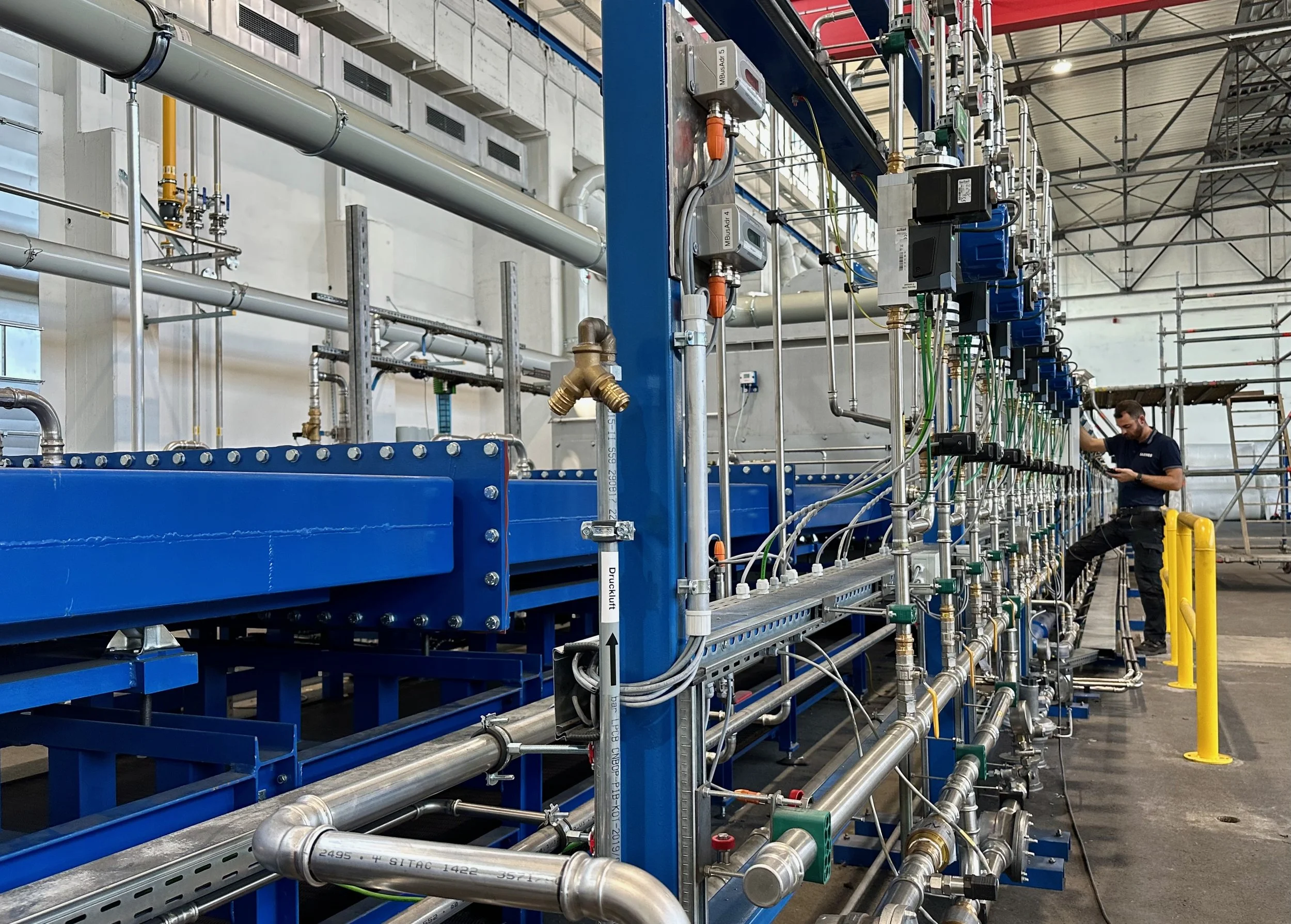 Industrielle Rohrleitungen und Ventile in einer Fabrikhalle, mit einem Arbeiter im Hintergrund, der ein Messgerät benutzt.